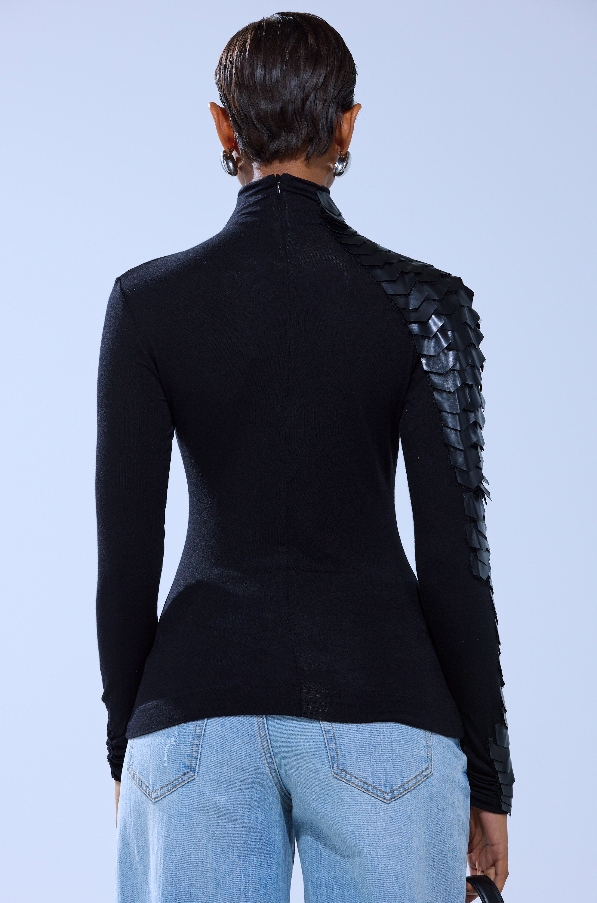 A person with slicked-back short hair stands facing away, wearing the NASTASIA SCALE TOP— a fitted black long-sleeve with geometric faux leather cutouts on one shoulder— paired with light blue jeans and holding a small black object.