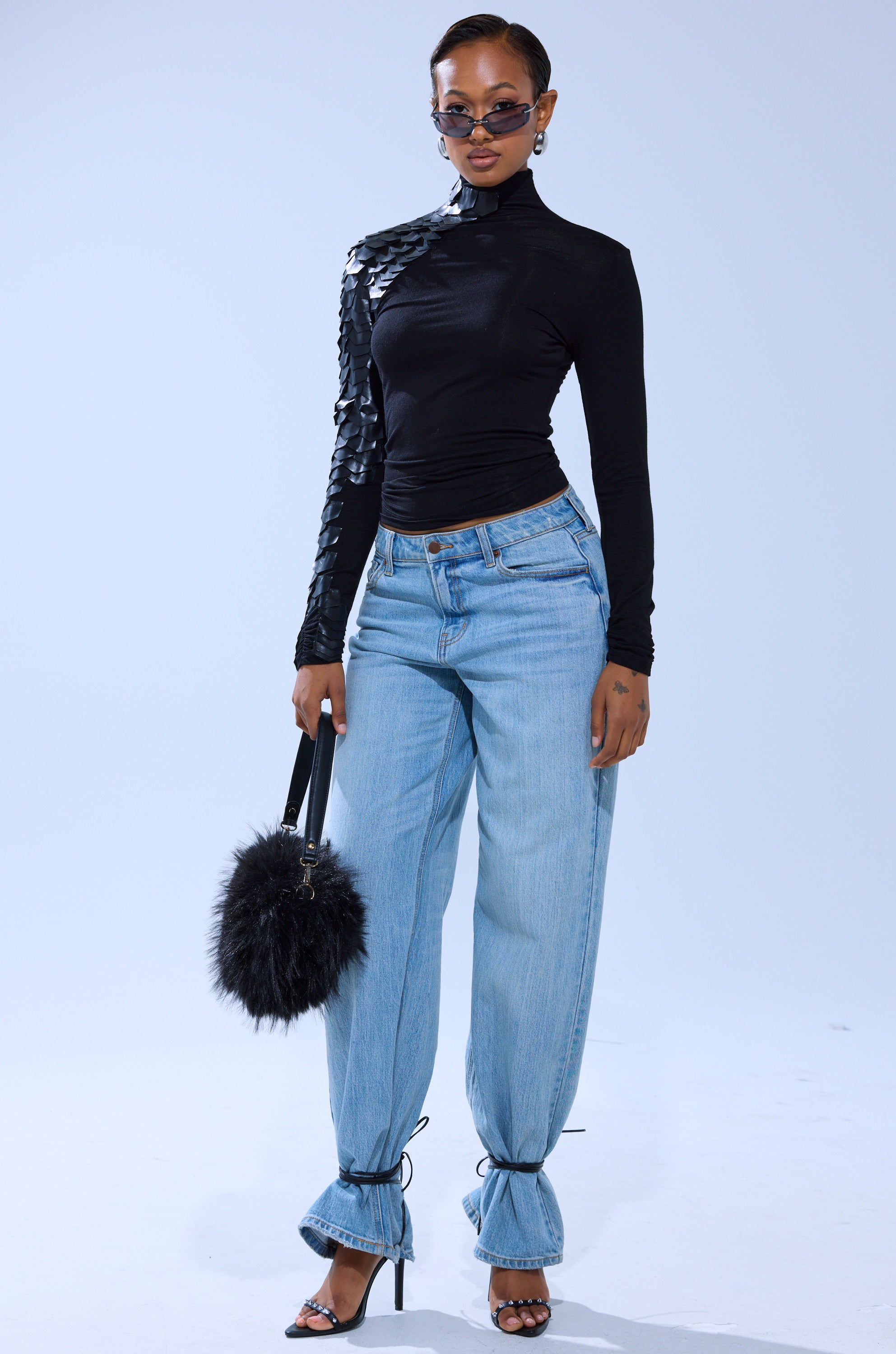 A woman wears the NASTASIA SCALE TOP—a fitted black turtleneck with geometric faux leather cutouts on one shoulder—paired with light blue high-waisted jeans, strappy black heels, sunglasses, and a fluffy black handbag.