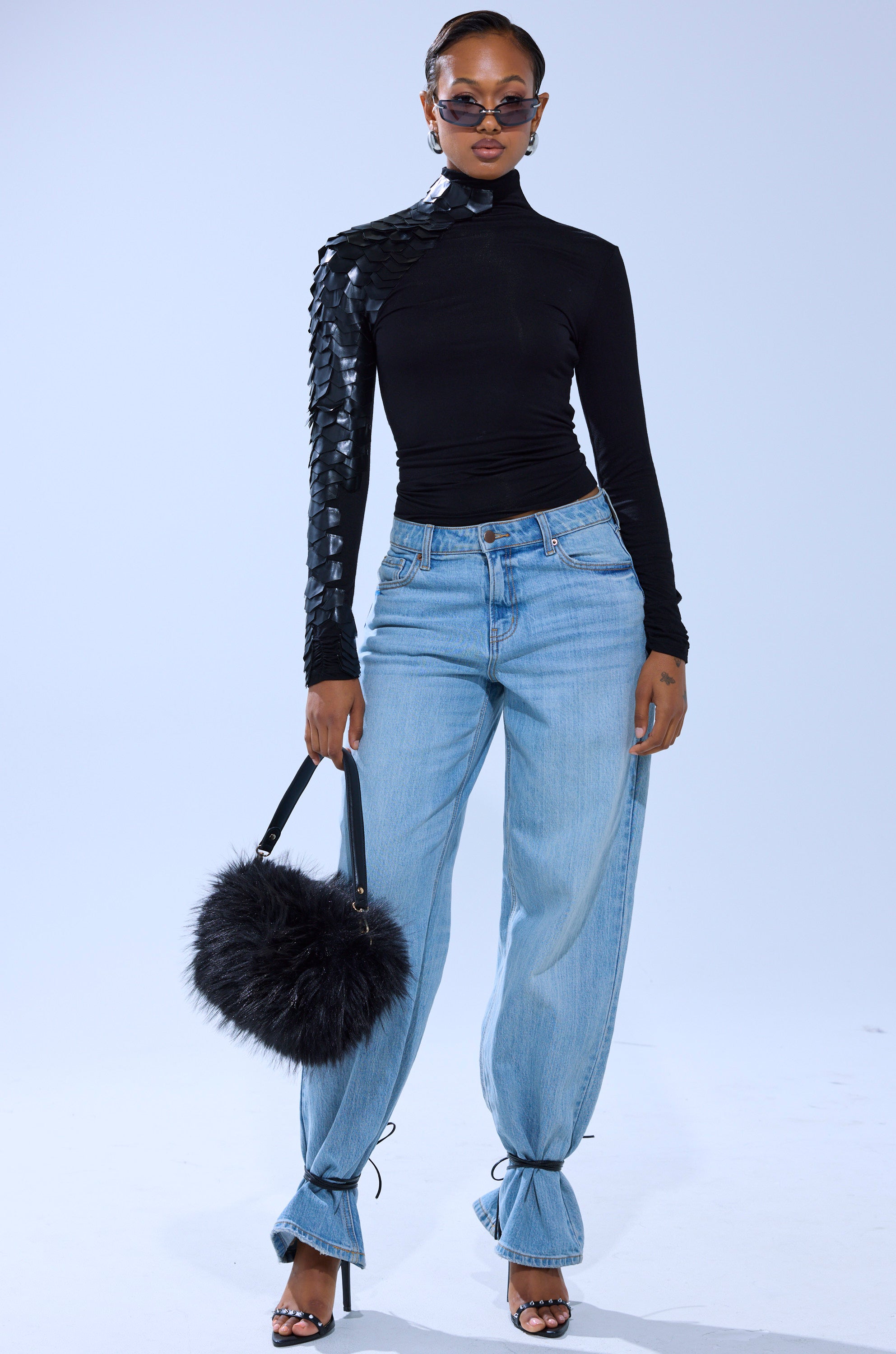 A stylish woman in a NASTASIA SCALE TOP with geometric faux leather cutouts, light blue loose-fit jeans, black heeled sandals, dark sunglasses, and holding a fluffy black handbag.