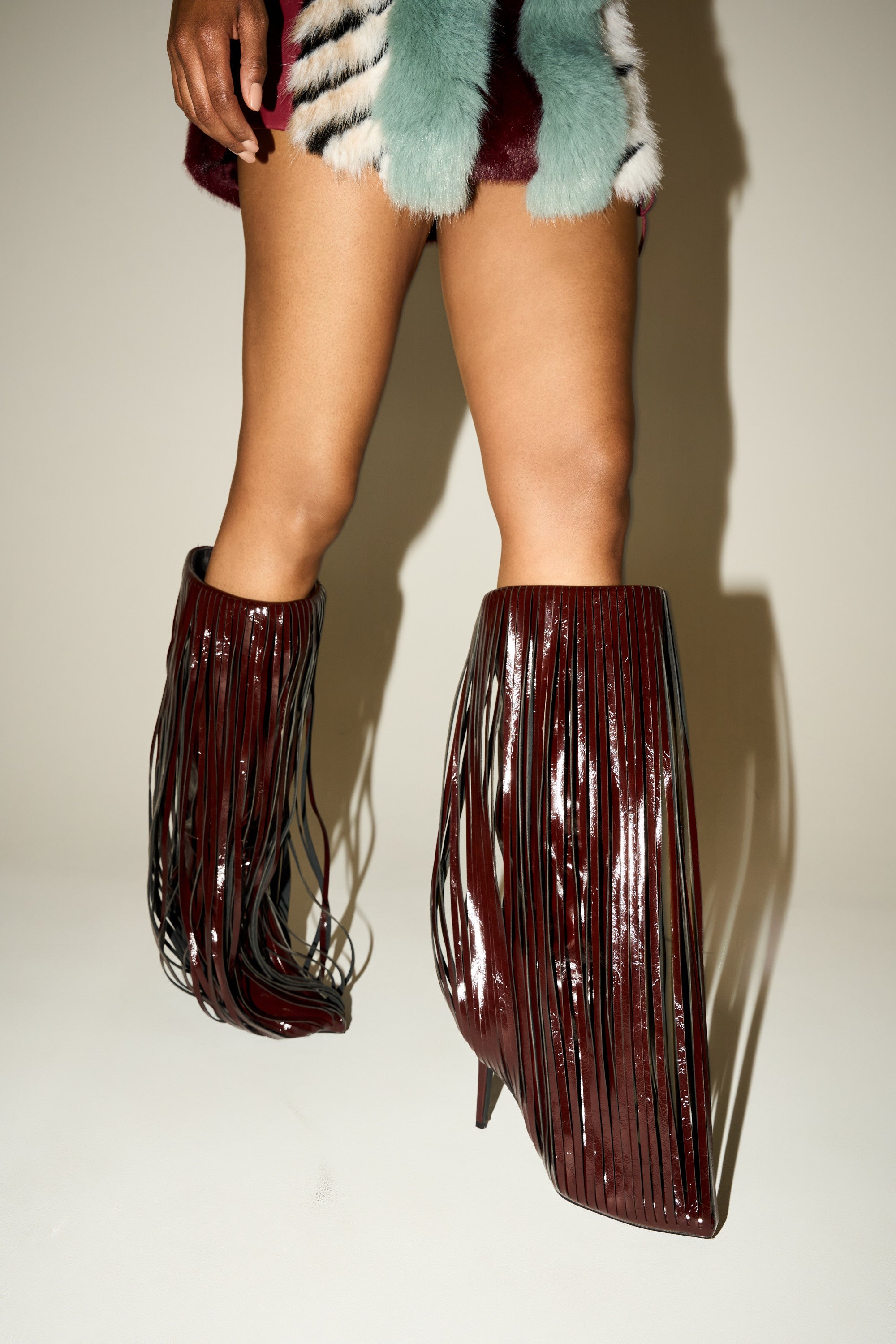Only their legs are visible against a plain background, highlighting the striking knee-high AZALEA WANG XANTARA FRINGE BURGUNDY STILETTO BOOT with long, thin fringe details, paired with a fur-trimmed skirt. 0