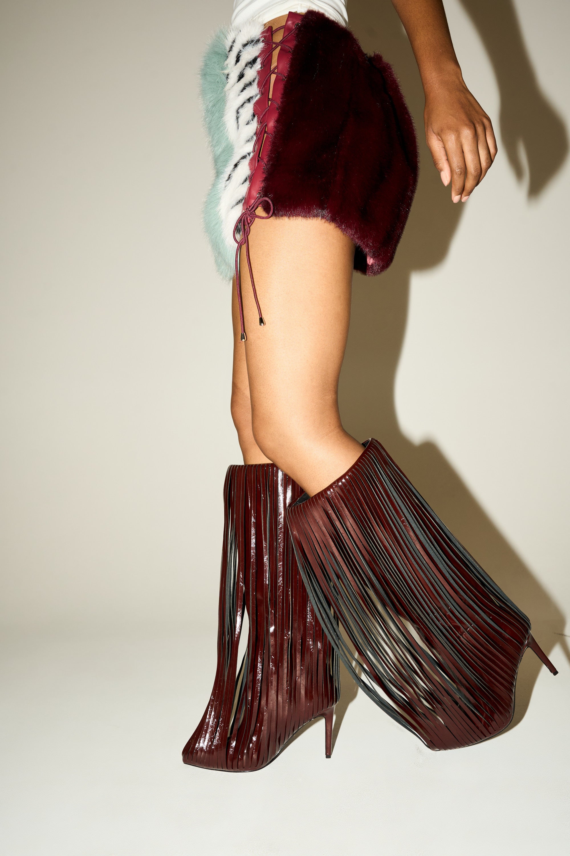 A person poses in a side profile, wearing furry lace-up shorts and the AZALEA WANG XANTARA FRINGE BURGUNDY STILETTO BOOT with shiny knee-high style and dramatic long fringe. The background is plain and light-colored. 2