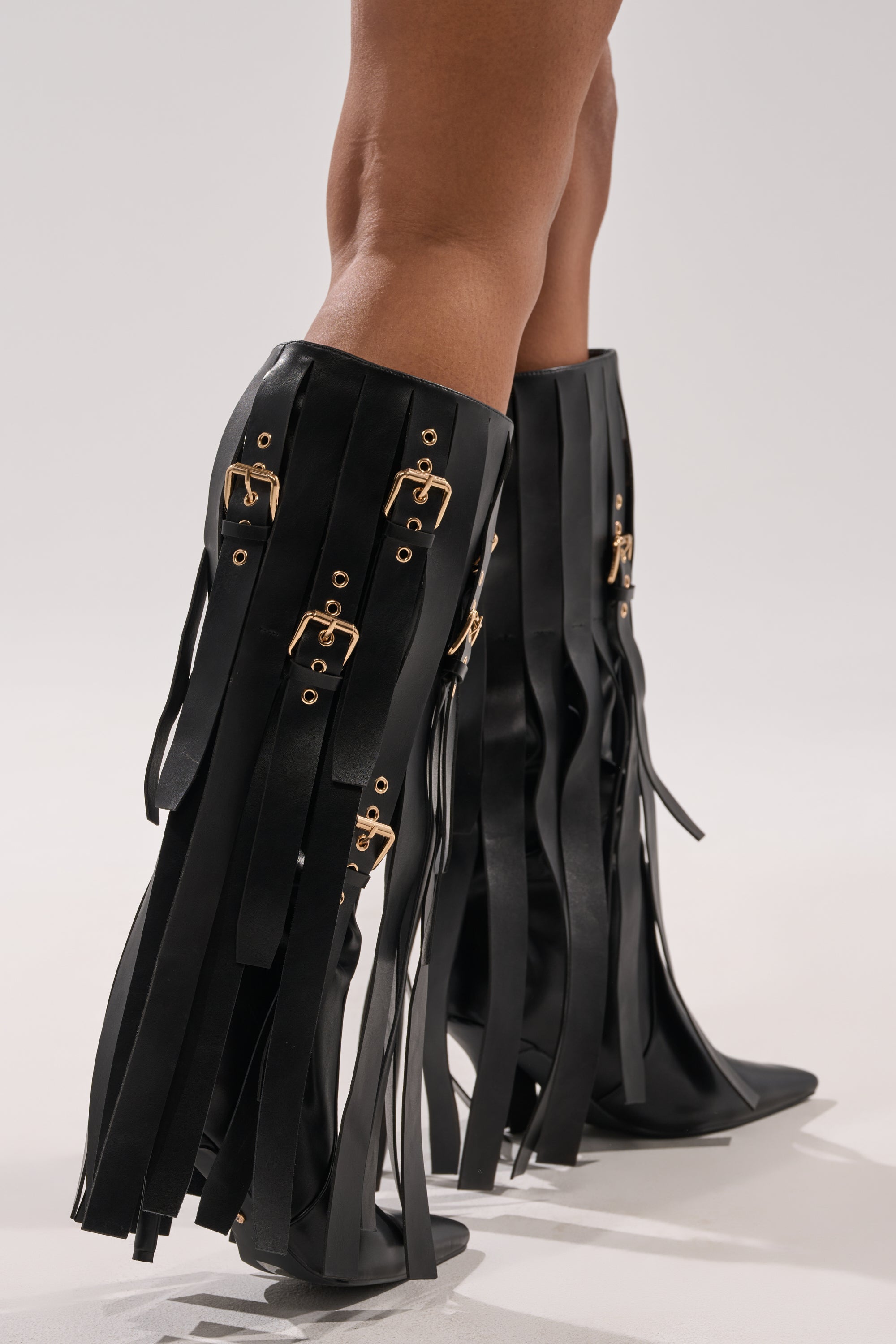 A person wearing the AZALEA WANG XANTHE BLACK BELT EMBELLISHED STILETO BOOT with knee-high design, long fringe straps, and gold buckle details stands on a light-colored surface.