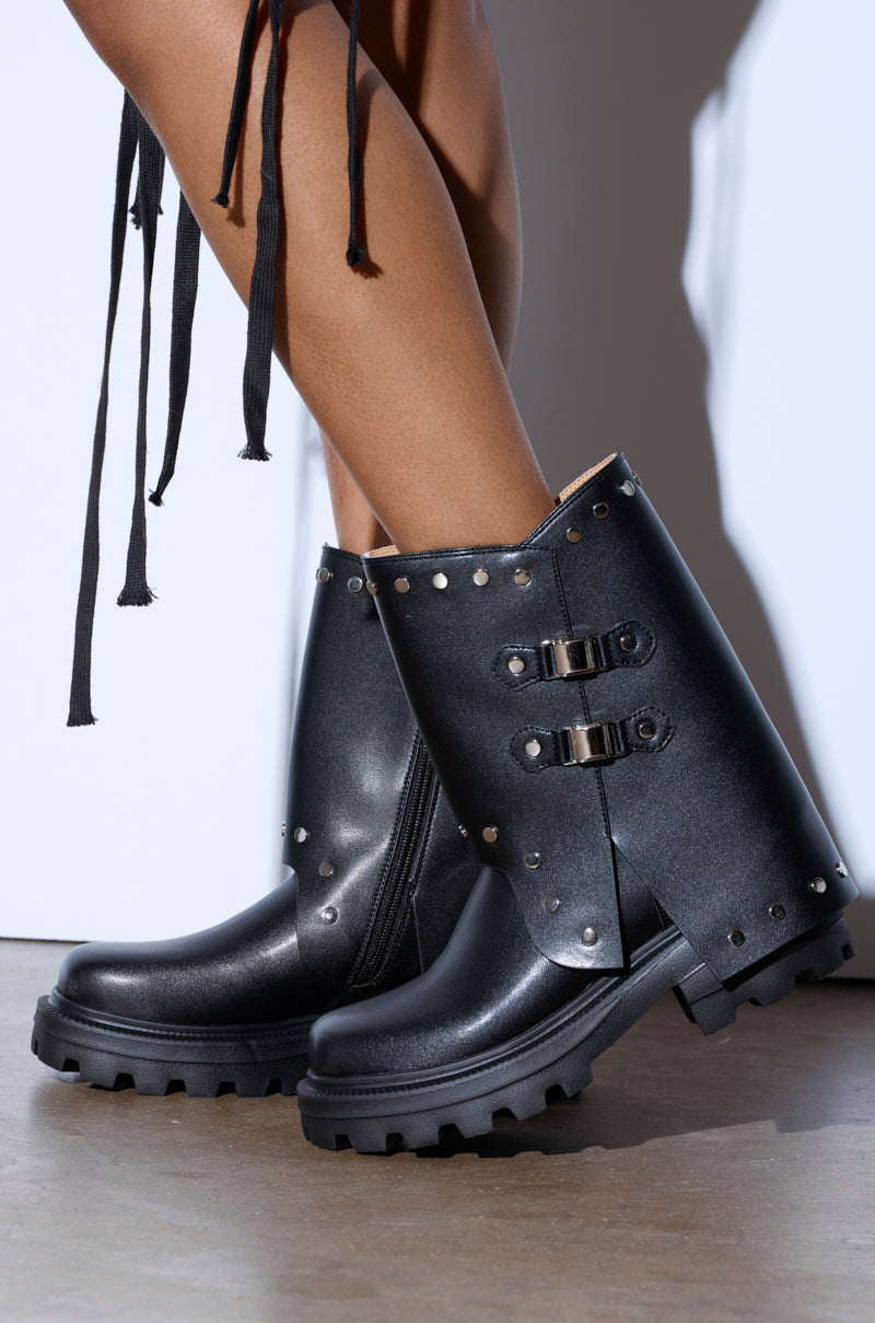 A close-up shows a person wearing the AZALEA WANG XAVIER BLACK WITH METAL FLAT BOOTIE, featuring rugged soles, decorative straps with metal buckles, metallic stud trims, and a small slit near the top. The person stands on a light-colored floor.