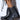 A close-up shows a person wearing the AZALEA WANG XAVIER BLACK WITH METAL FLAT BOOTIE, featuring rugged soles, decorative straps with metal buckles, metallic stud trims, and a small slit near the top. The person stands on a light-colored floor.
