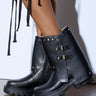 A close-up shows a person wearing the AZALEA WANG XAVIER BLACK WITH METAL FLAT BOOTIE, featuring rugged soles, decorative straps with metal buckles, metallic stud trims, and a small slit near the top. The person stands on a light-colored floor.