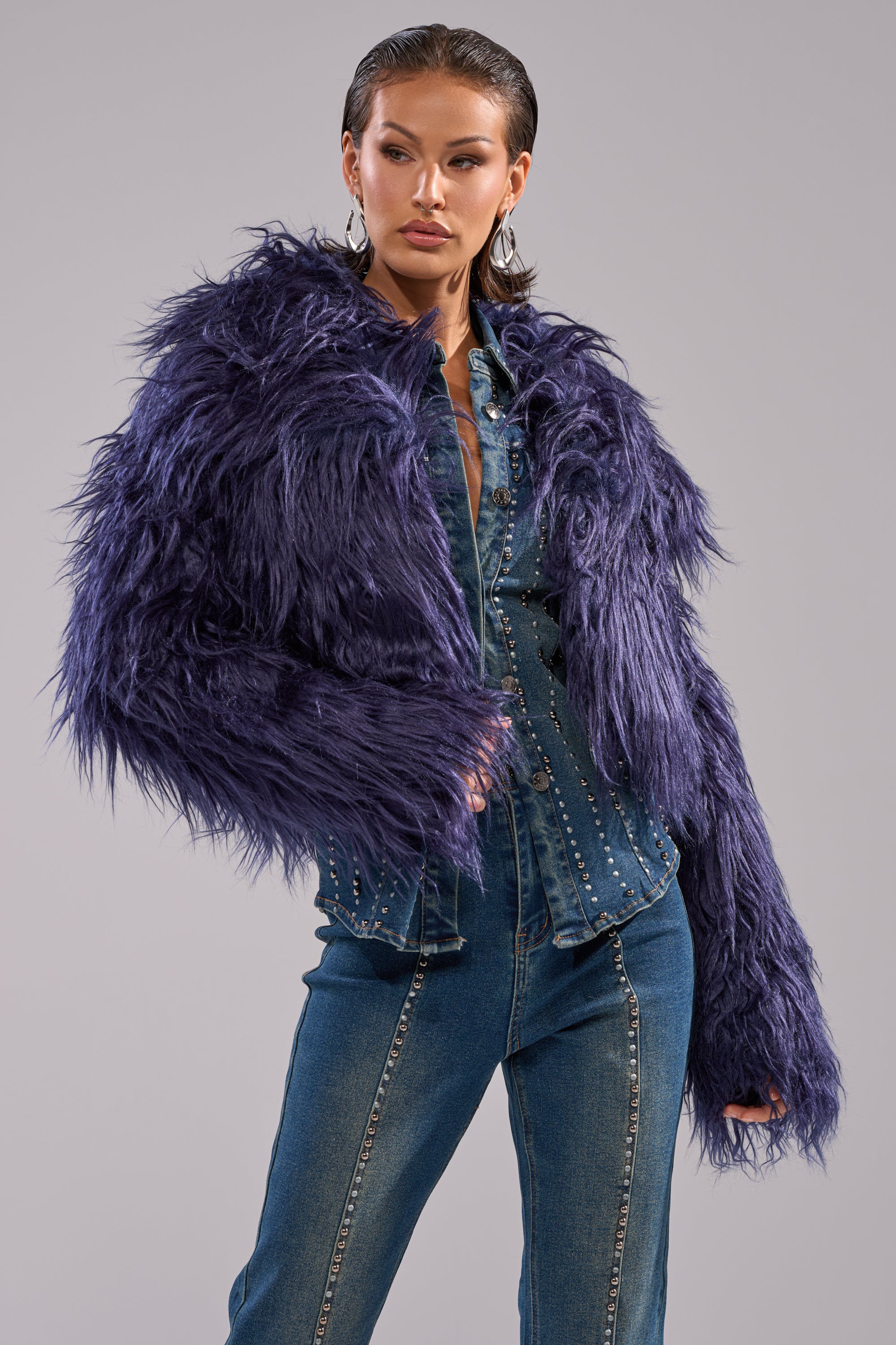 A woman poses confidently against a light gray background wearing the LIFE OF THE PARTY FAUX FUR, a textured, oversized purple jacket layered over a studded denim jumpsuit.