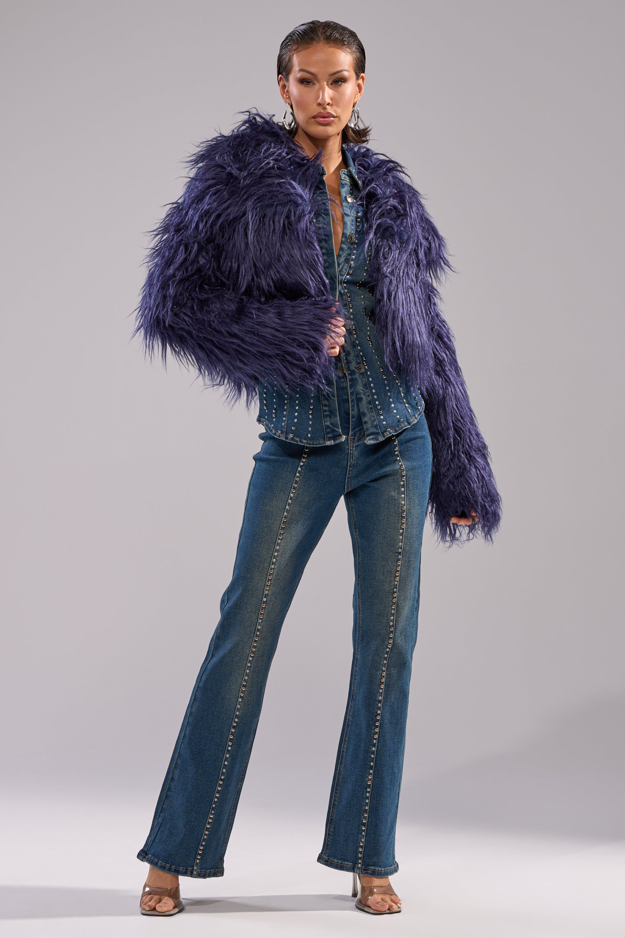 A woman poses confidently in the LIFE OF THE PARTY FAUX FUR jacket over a studded denim shirt and flared blue jeans with visible seams, wearing high-heeled sandals against a plain gray background.