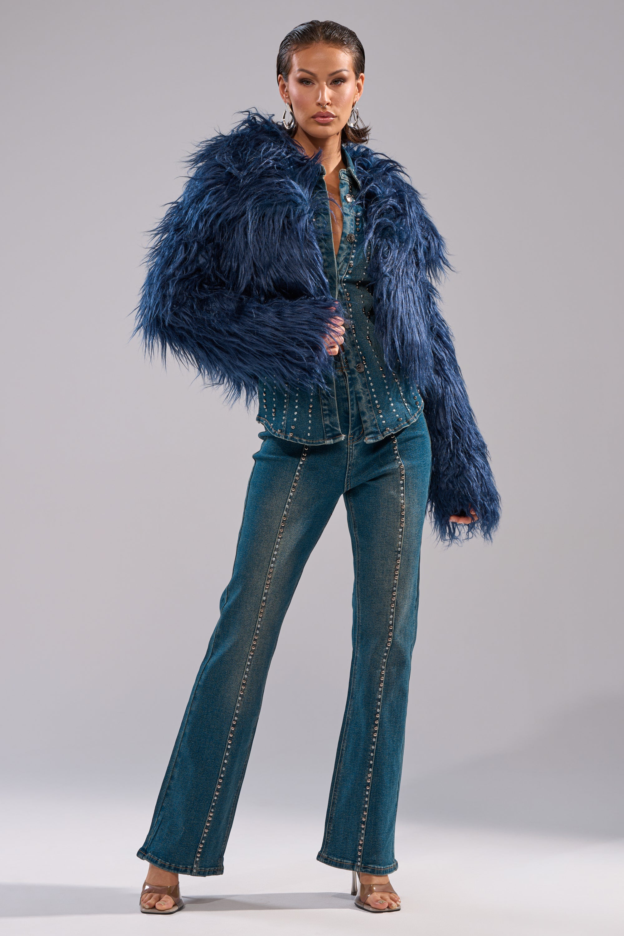 A person models the LIFE OF THE PARTY FAUX FUR jacket over a studded denim shirt and flared jeans with silver details, styled with hoop earrings and heeled sandals against a plain light gray background. 9