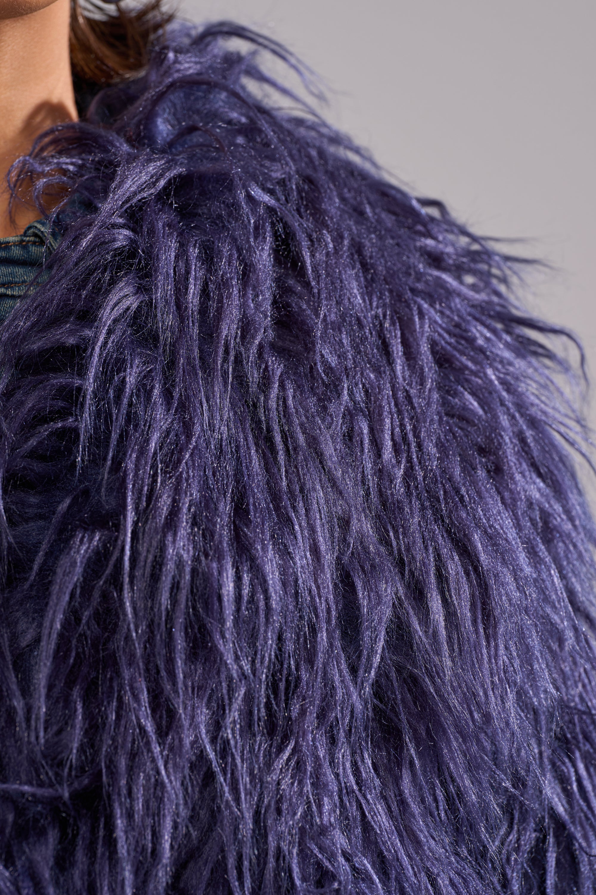 Close-up of a person wearing the LIFE OF THE PARTY FAUX FUR, a deep purple cropped coat that highlights the texture and shine of its long, fluffy fibers against a neutral, softly blurred background.