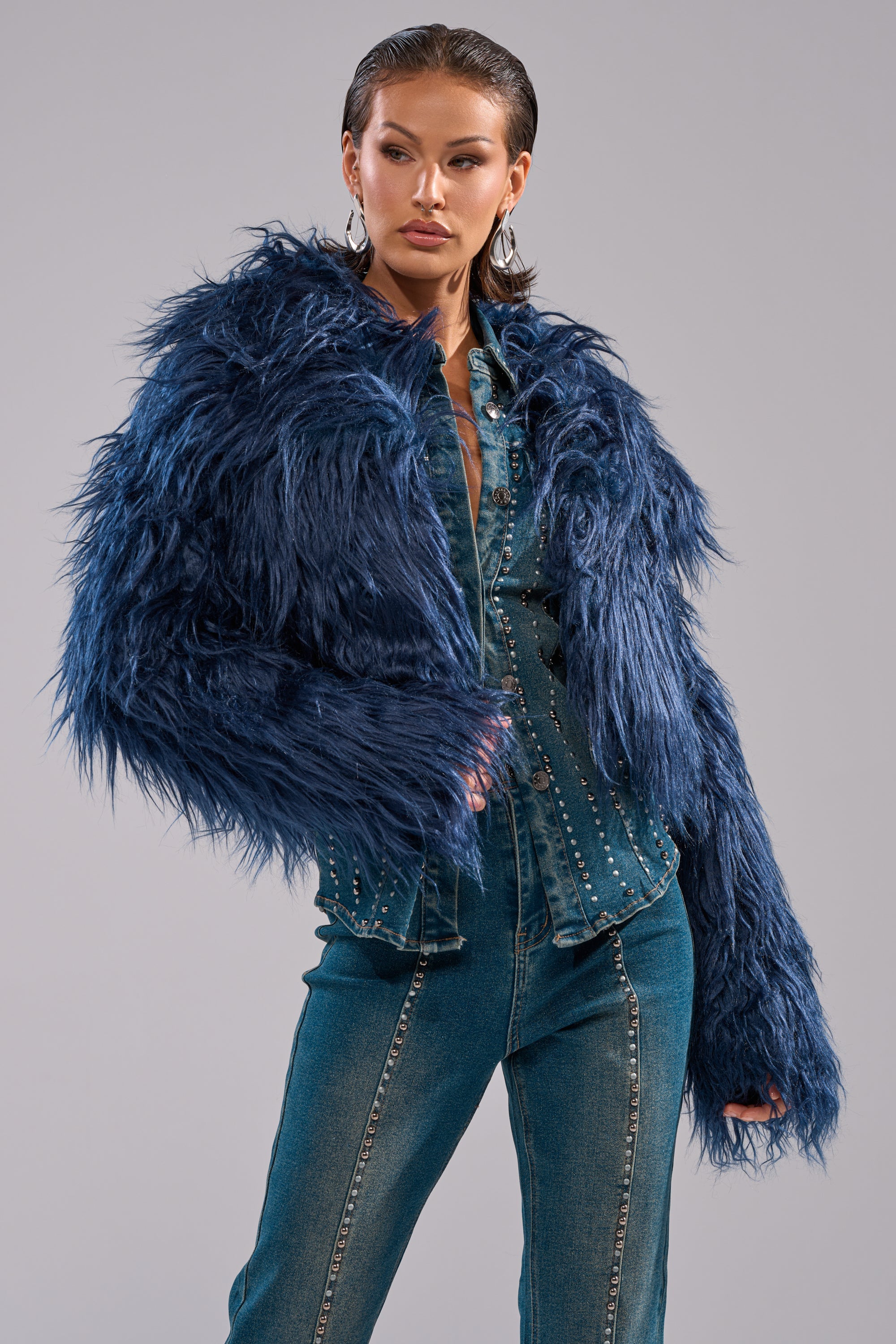 A woman poses confidently against a plain gray background, wearing the LIFE OF THE PARTY FAUX FUR coat over a studded denim jumpsuit. 0