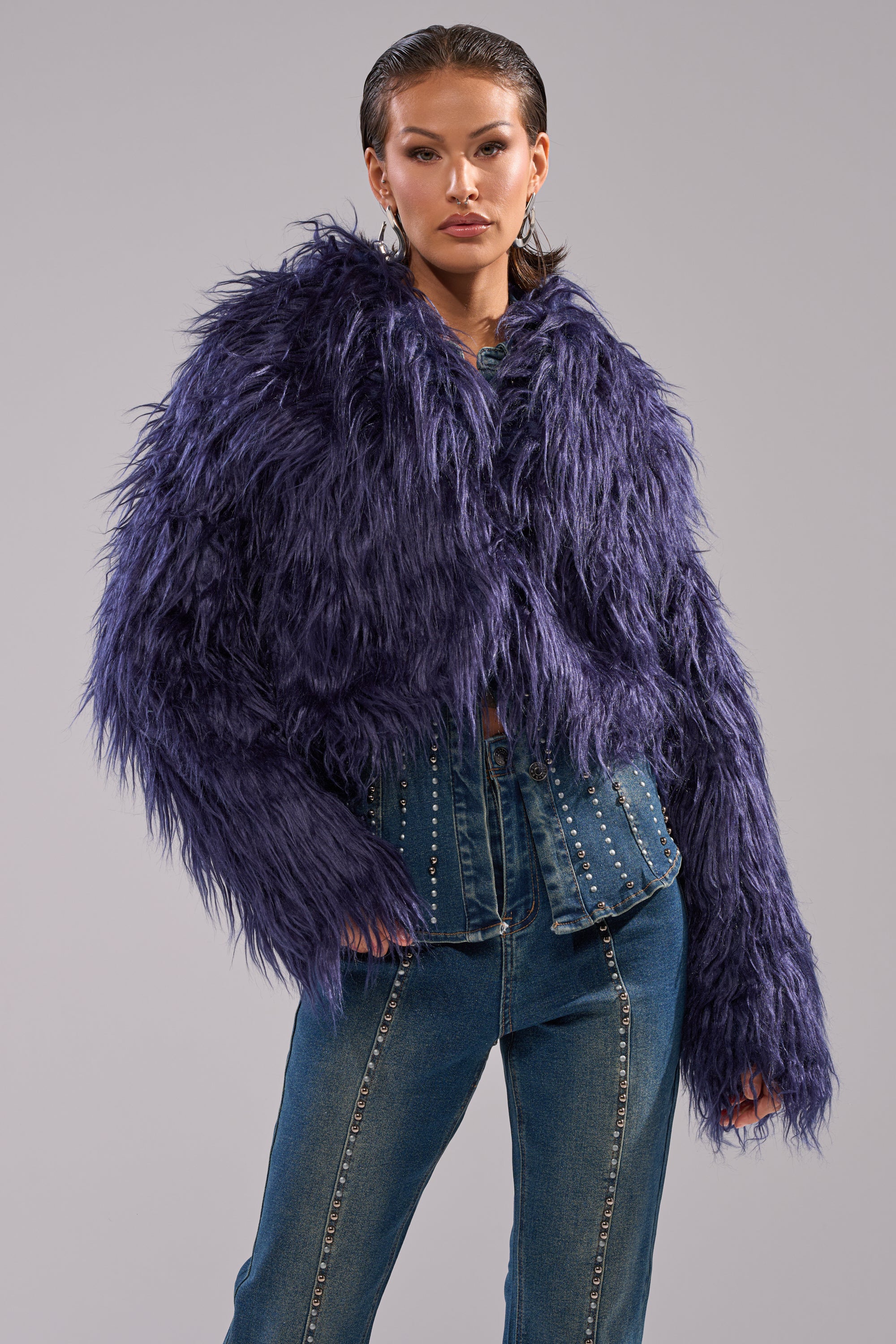 A person models the LIFE OF THE PARTY FAUX FUR—a shaggy purple jacket—layered over a studded denim shirt and blue jeans, standing confidently against a plain grey background.