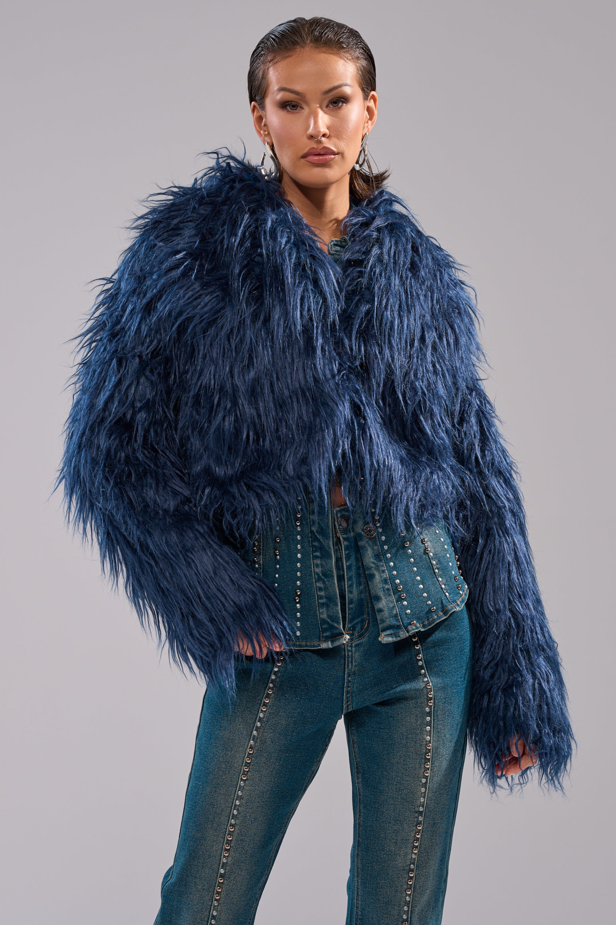 A person wears the LIFE OF THE PARTY FAUX FUR, a cropped navy blue faux fur jacket, paired with high-waisted blue jeans with decorative studs, standing against a plain gray background. 2