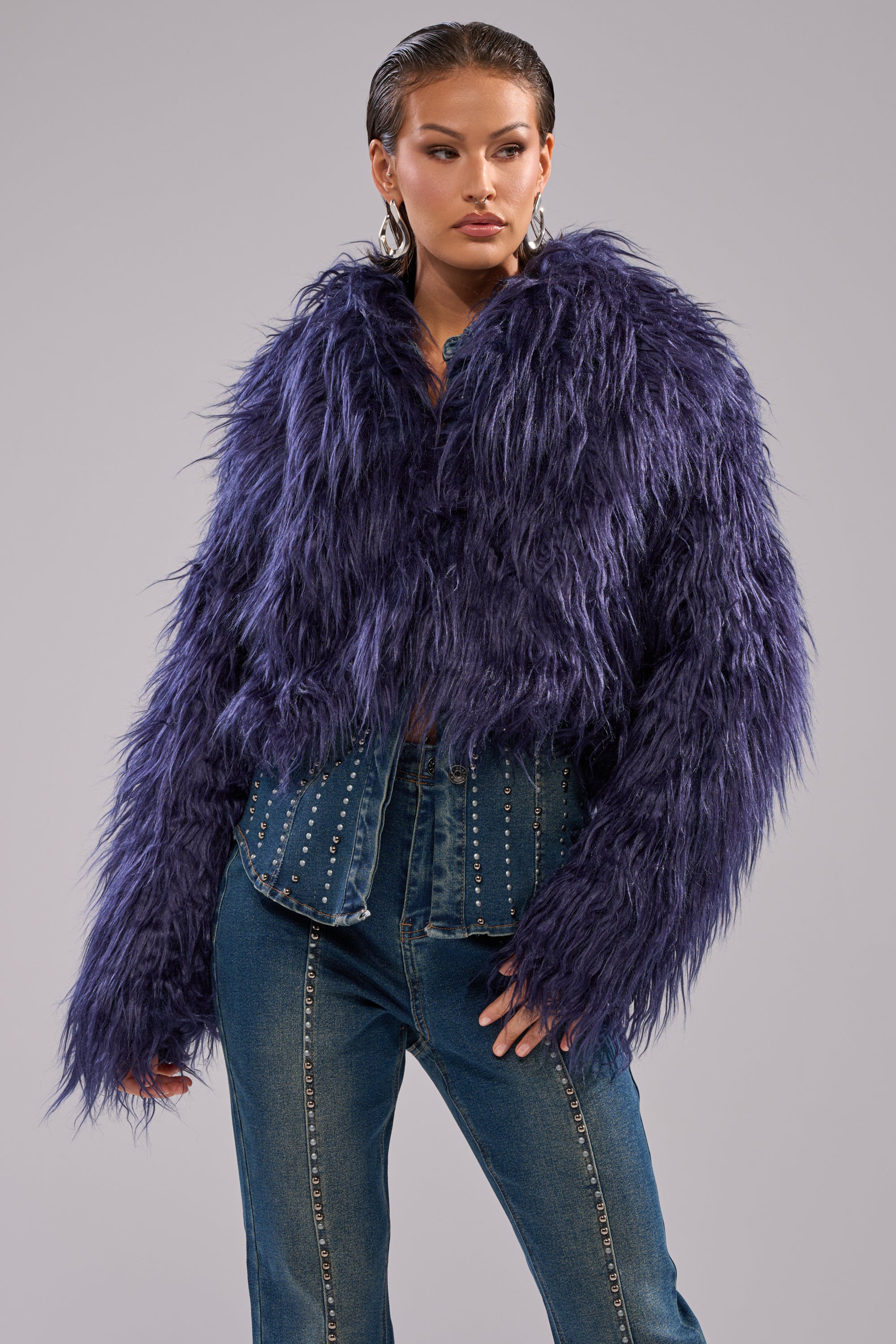 A person wears the LIFE OF THE PARTY FAUX FUR jacket in fluffy purple over a studded denim shirt and blue jeans, standing against a plain light gray background.