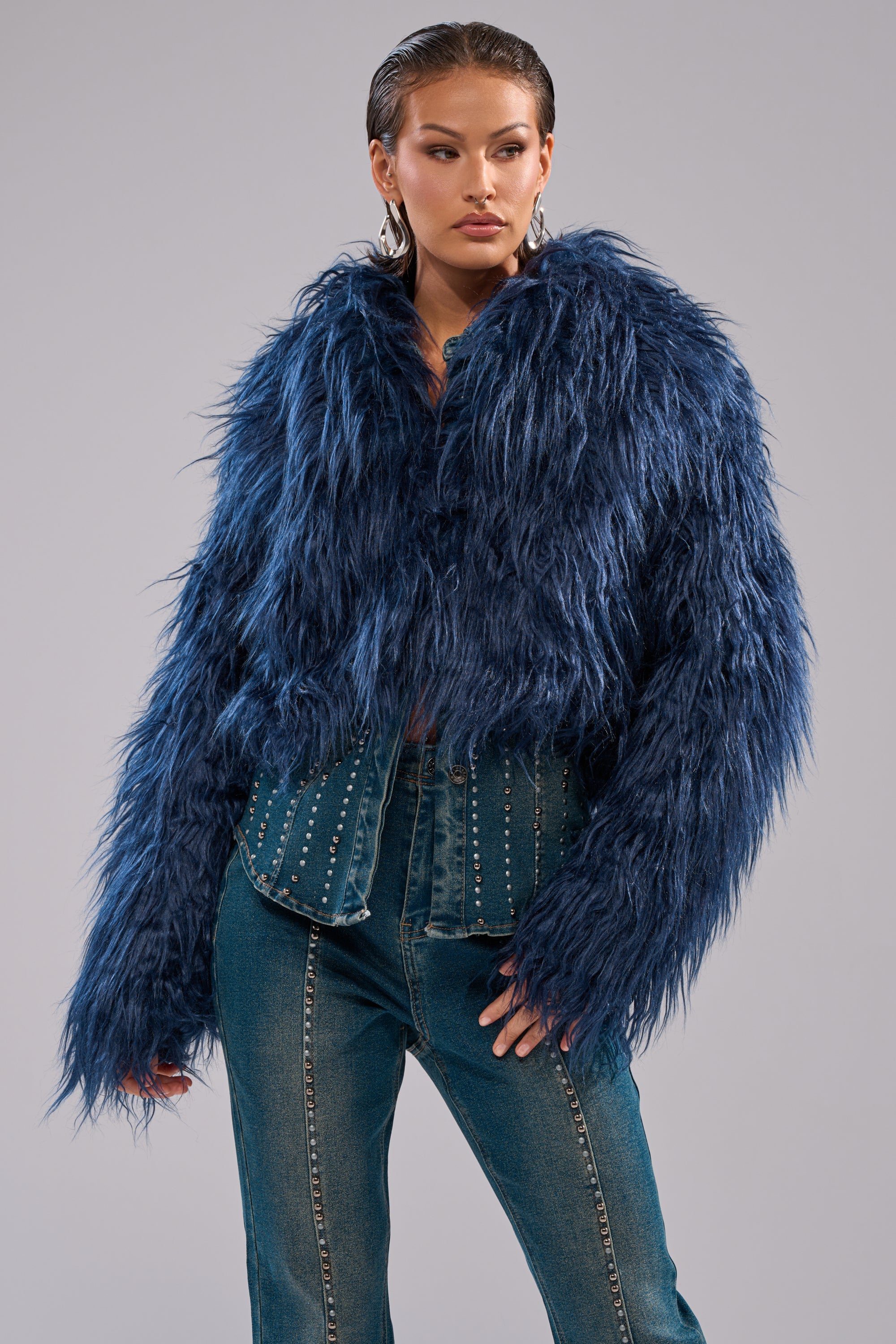 A person wears the LIFE OF THE PARTY FAUX FUR, a textured, oversized navy blue jacket, paired with high-waisted blue jeans with metallic studs, posing confidently against a plain light gray background. 3