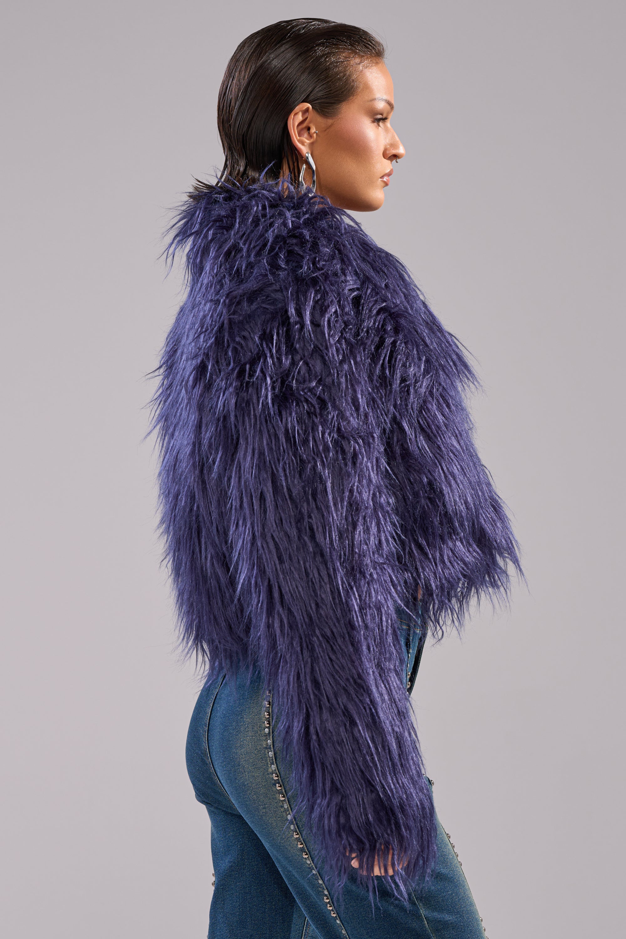 A woman with slicked-back hair wears the LIFE OF THE PARTY FAUX FUR, a shaggy, cropped purple jacket, and high-waisted blue jeans. She stands sideways against a plain light gray background.