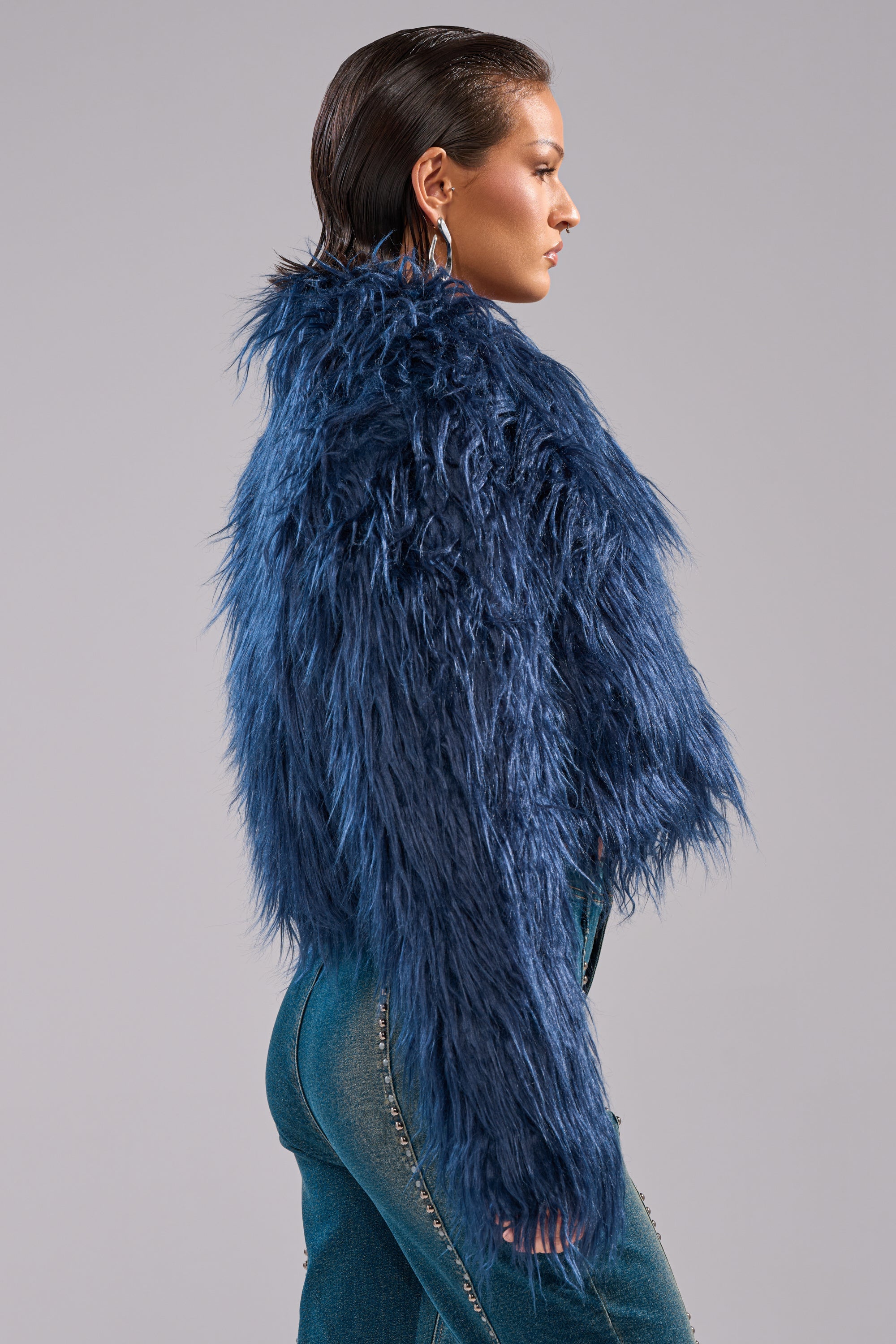 A person with slicked-back hair stands sideways against a plain gray background, wearing the blue LIFE OF THE PARTY FAUX FUR jacket and jeans adorned with silver studs. 5
