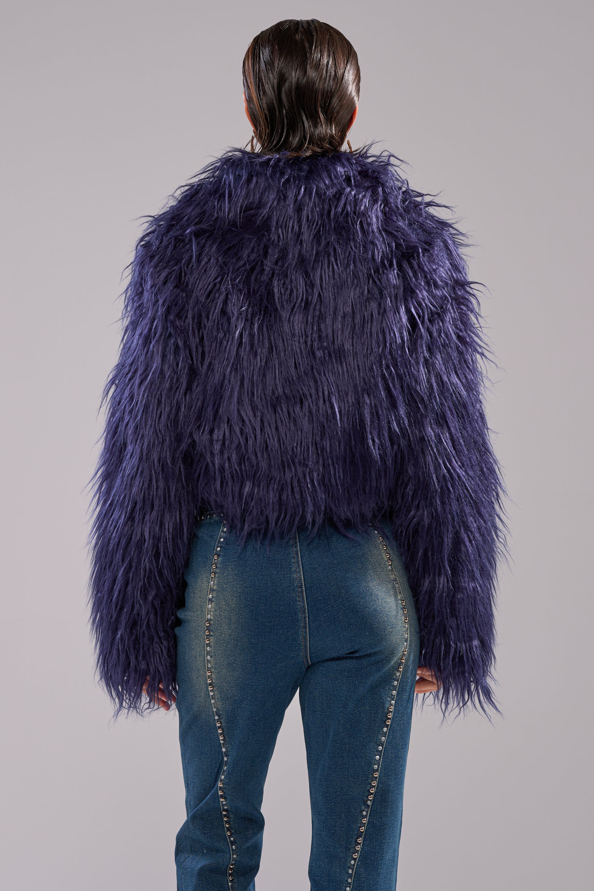 A person faces away from the camera, wearing the LIFE OF THE PARTY FAUX FUR—a shaggy, dark purple cropped jacket—paired with blue jeans featuring decorative stitching against a plain light gray background.