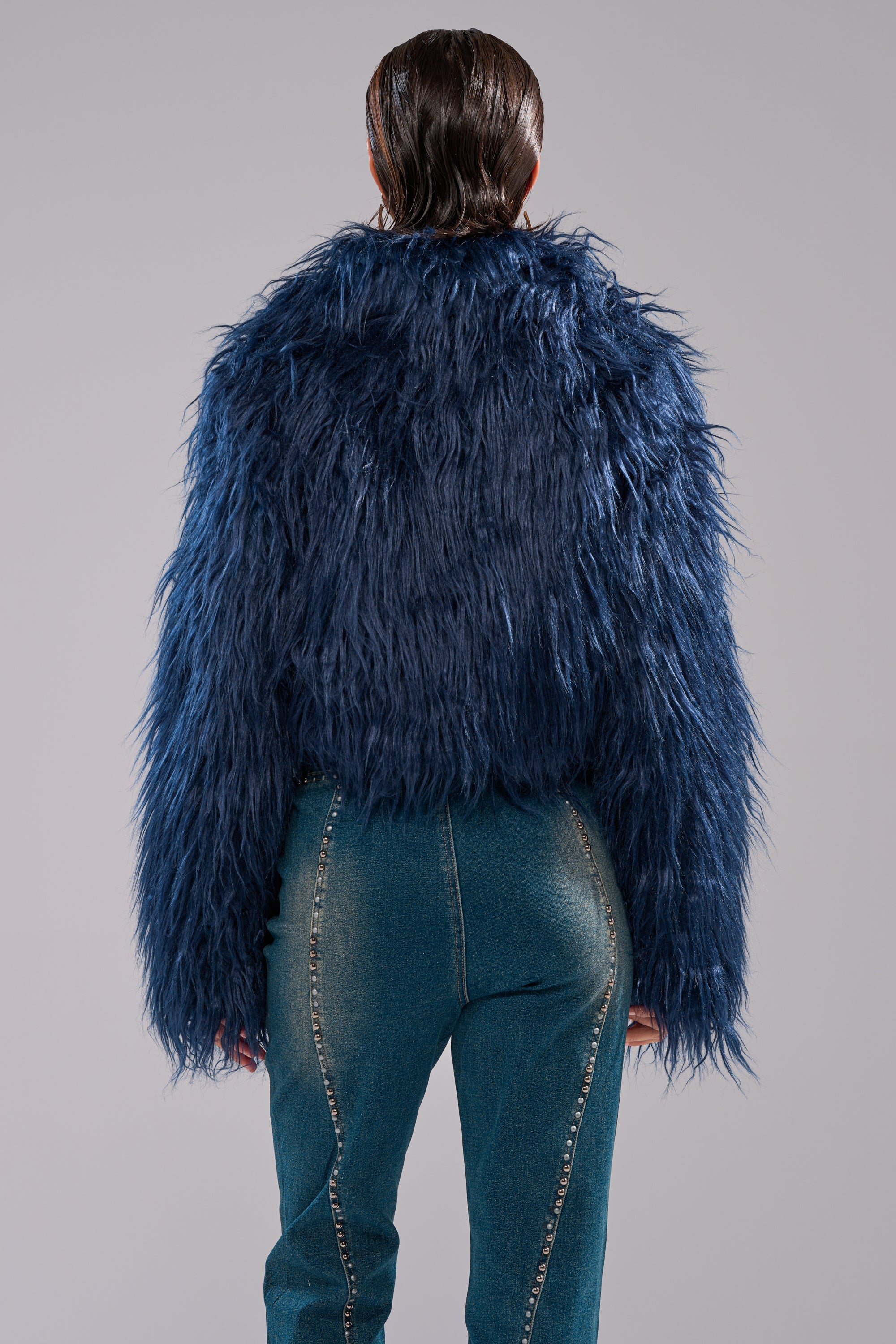 A person faces away from the camera, wearing the LIFE OF THE PARTY FAUX FUR jacket in dark blue and high-waisted blue denim jeans with decorative stitching, set against a plain light gray background. 6