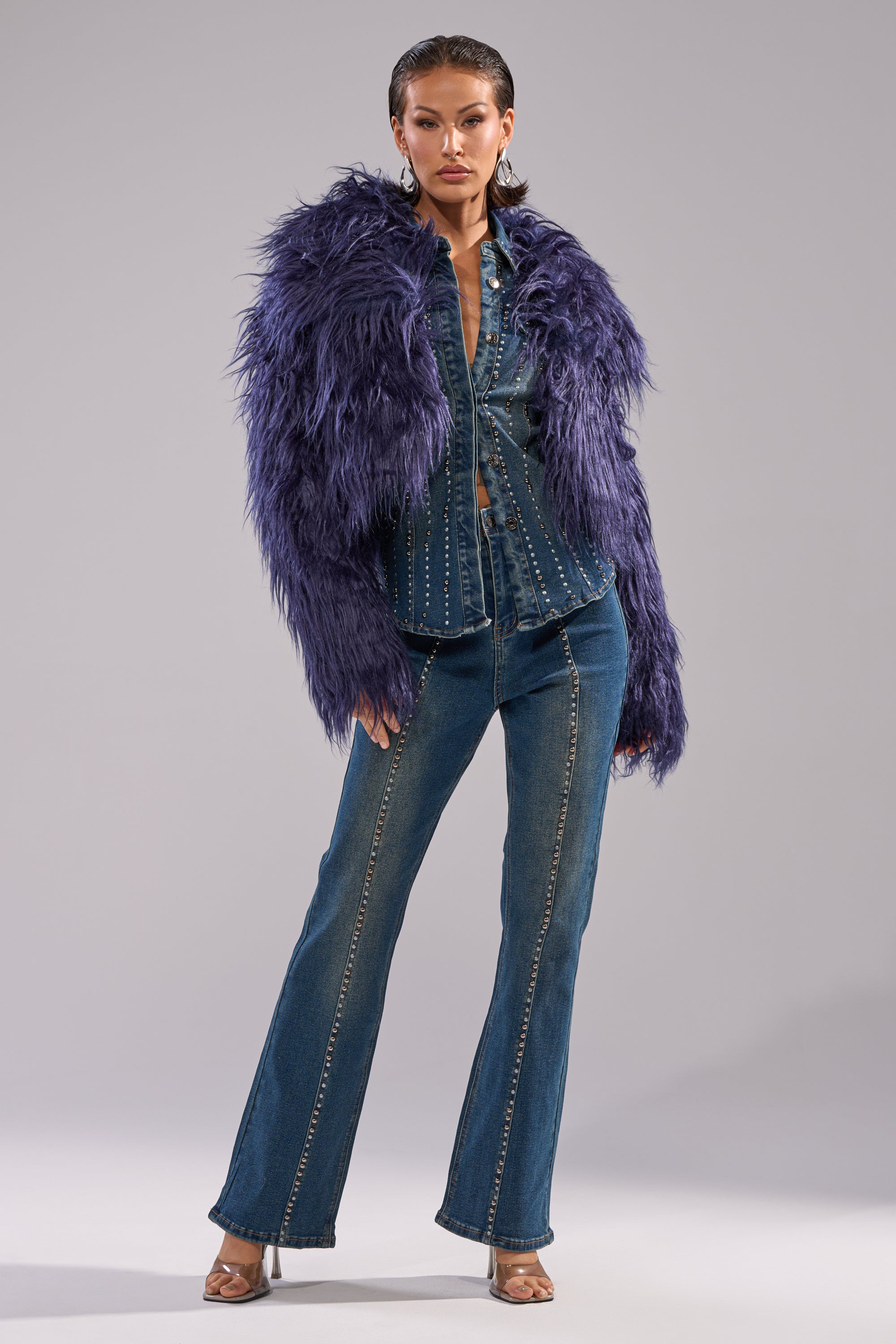 A woman stands confidently in the LIFE OF THE PARTY FAUX FUR jacket over a studded denim shirt and flared jeans with visible seams, paired with open-toe heels and hoop earrings against a plain background.