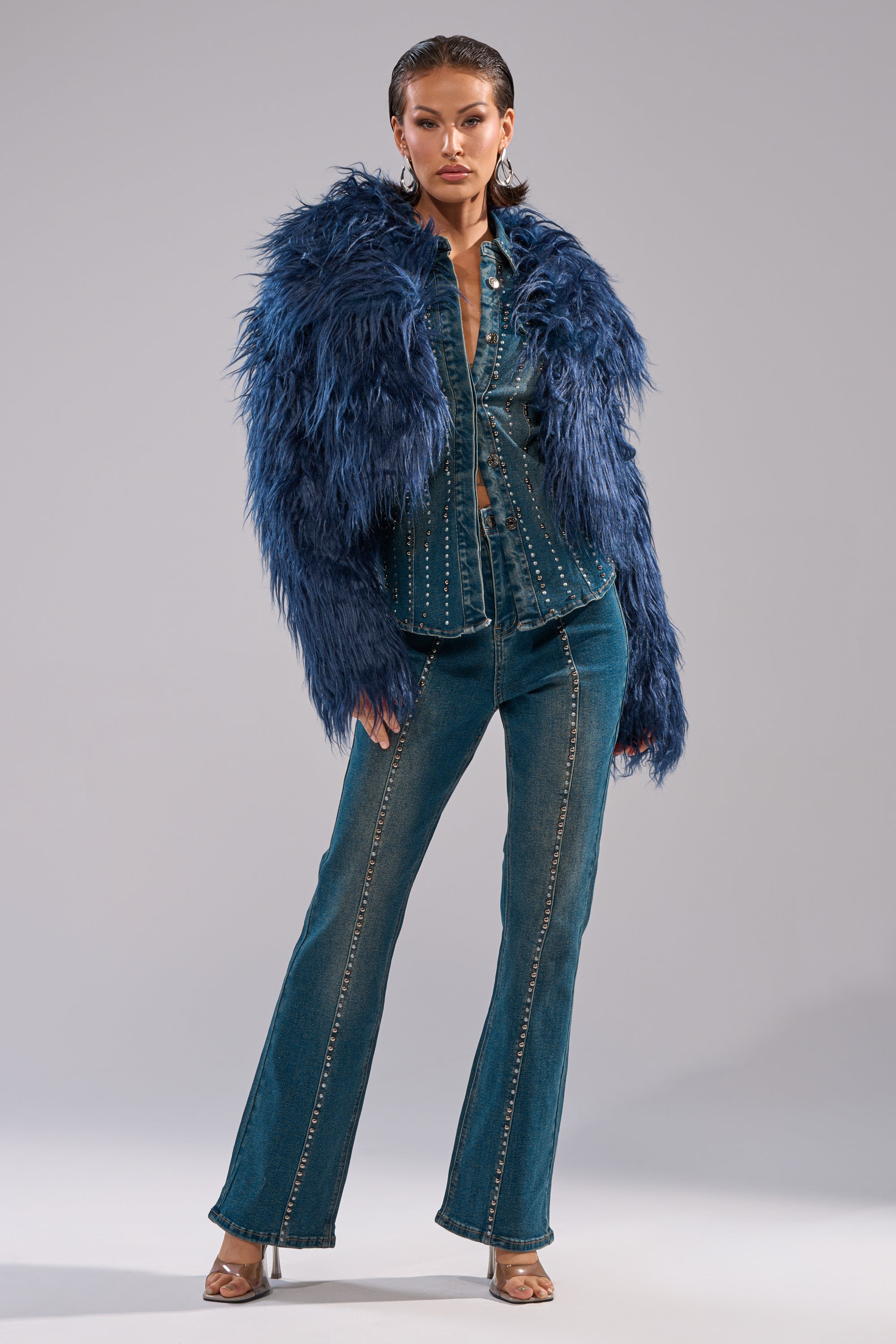 A person poses confidently in the LIFE OF THE PARTY FAUX FUR jacket over a studded denim shirt and flared jeans, accessorized with silver hoop earrings and open-toe heels, set against a plain light gray background. 7