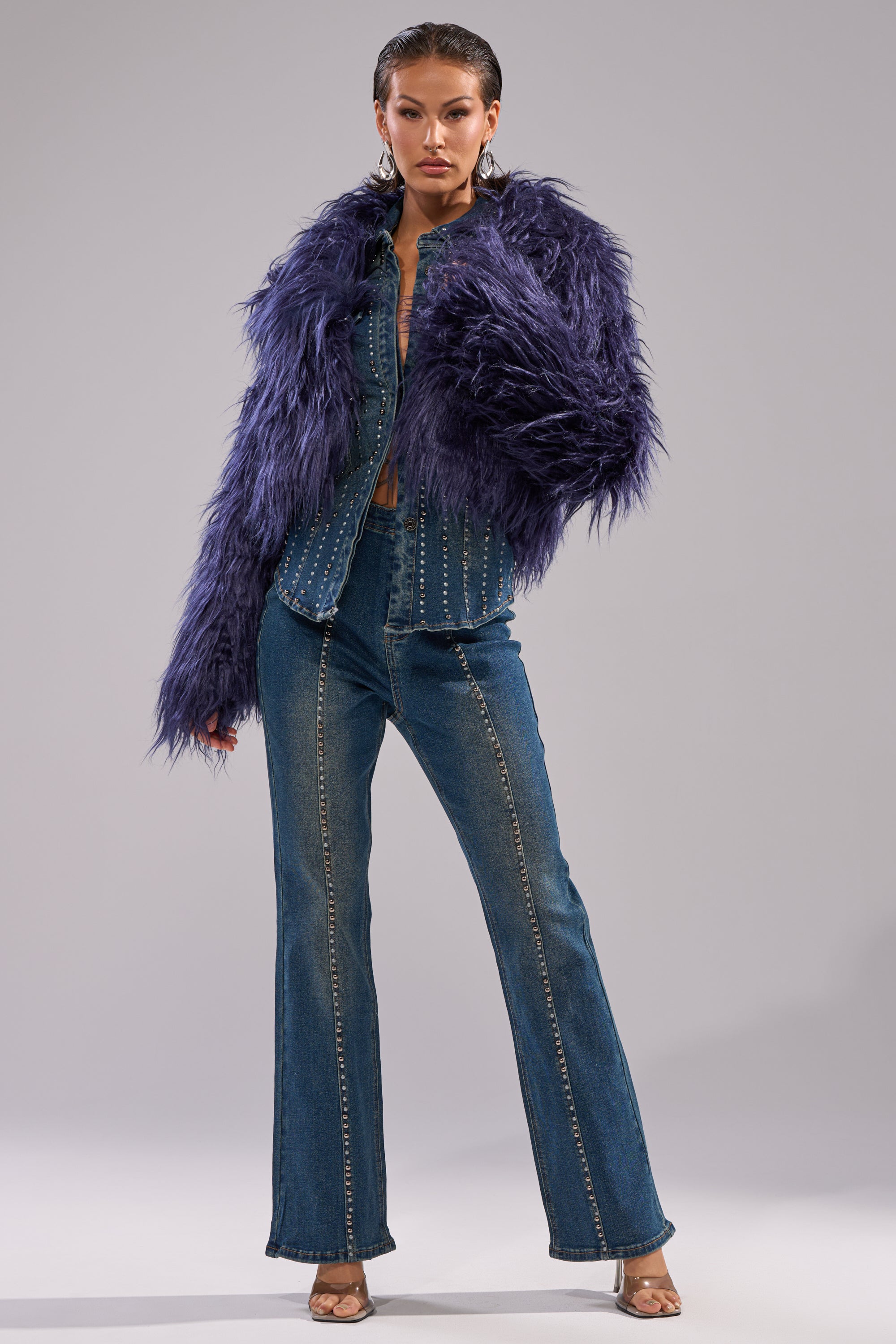 A woman poses confidently in the LIFE OF THE PARTY FAUX FUR jacket over a studded denim jumpsuit with flared legs, hoop earrings, and heeled sandals, standing against a plain gray background.