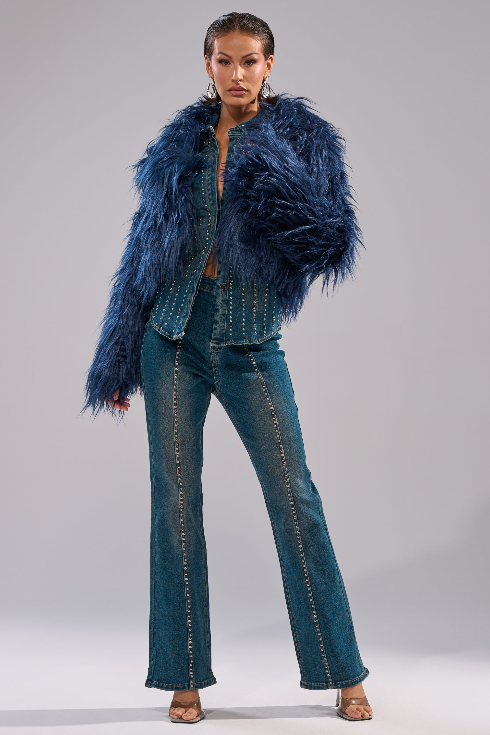 A woman stands confidently in the LIFE OF THE PARTY FAUX FUR jacket, paired with a studded, flared denim jumpsuit, open-toed heels, and statement earrings against a plain gray backdrop. 8