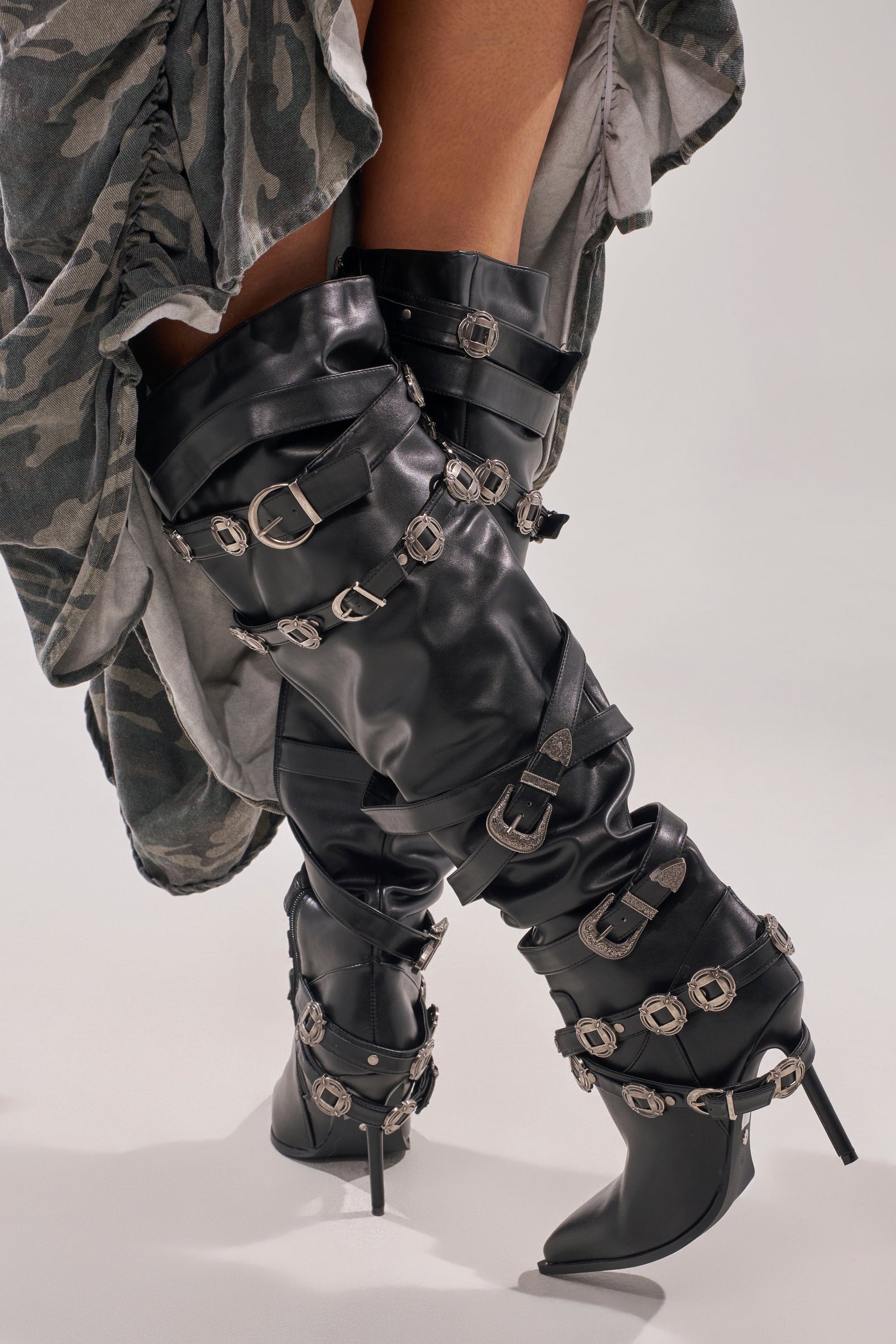 A person wearing AKIRA GOLD XERXES BLACK STILETTO METAL EMBELLISHED BOOTS, styled with a flowing camouflage-patterned skirt. 0