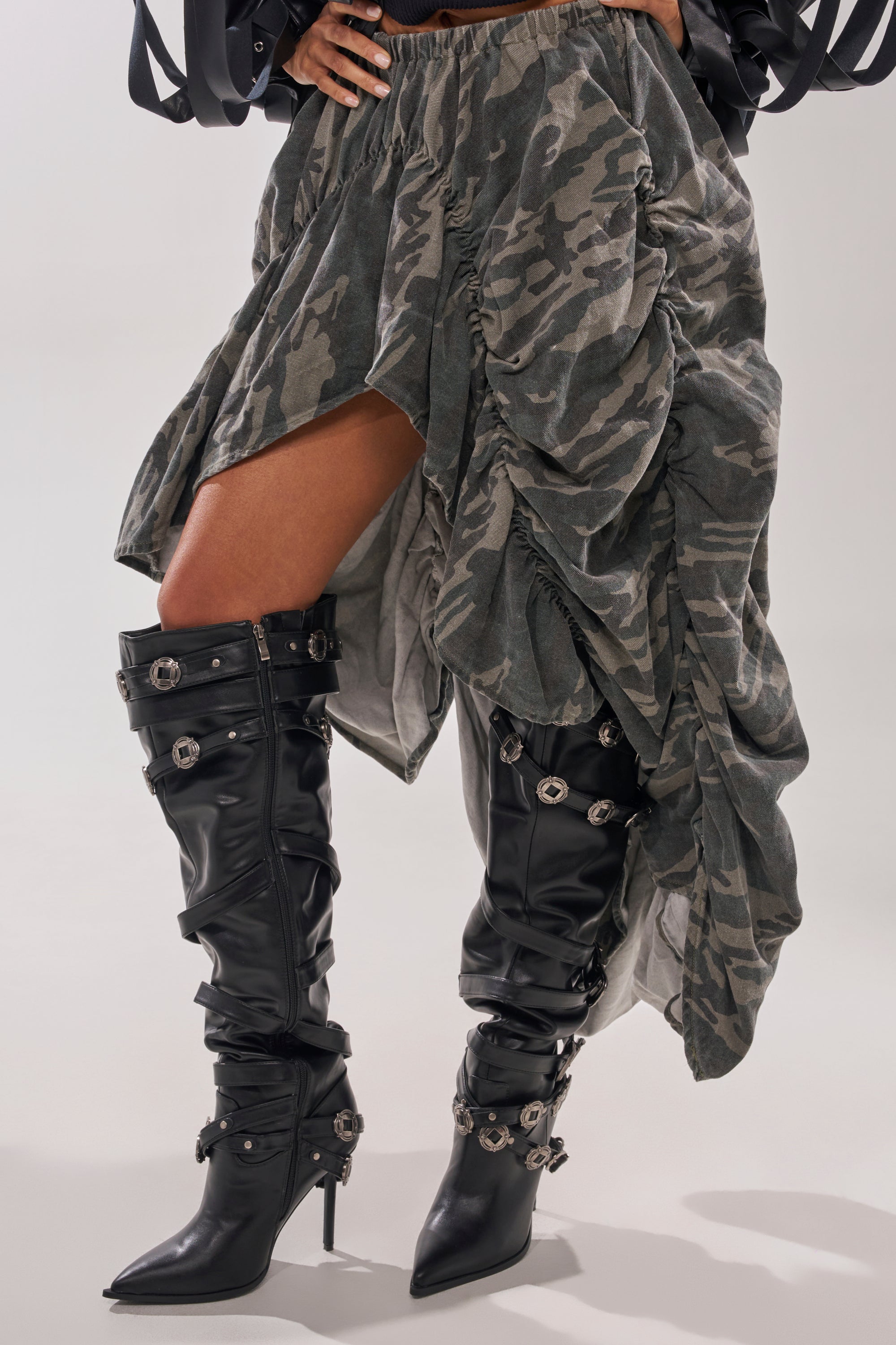 A person wears a camo-patterned ruched asymmetrical skirt with black AKIRA Gold Xerxes Stiletto Metal Embellished Boots. The upper body is out of frame. 1