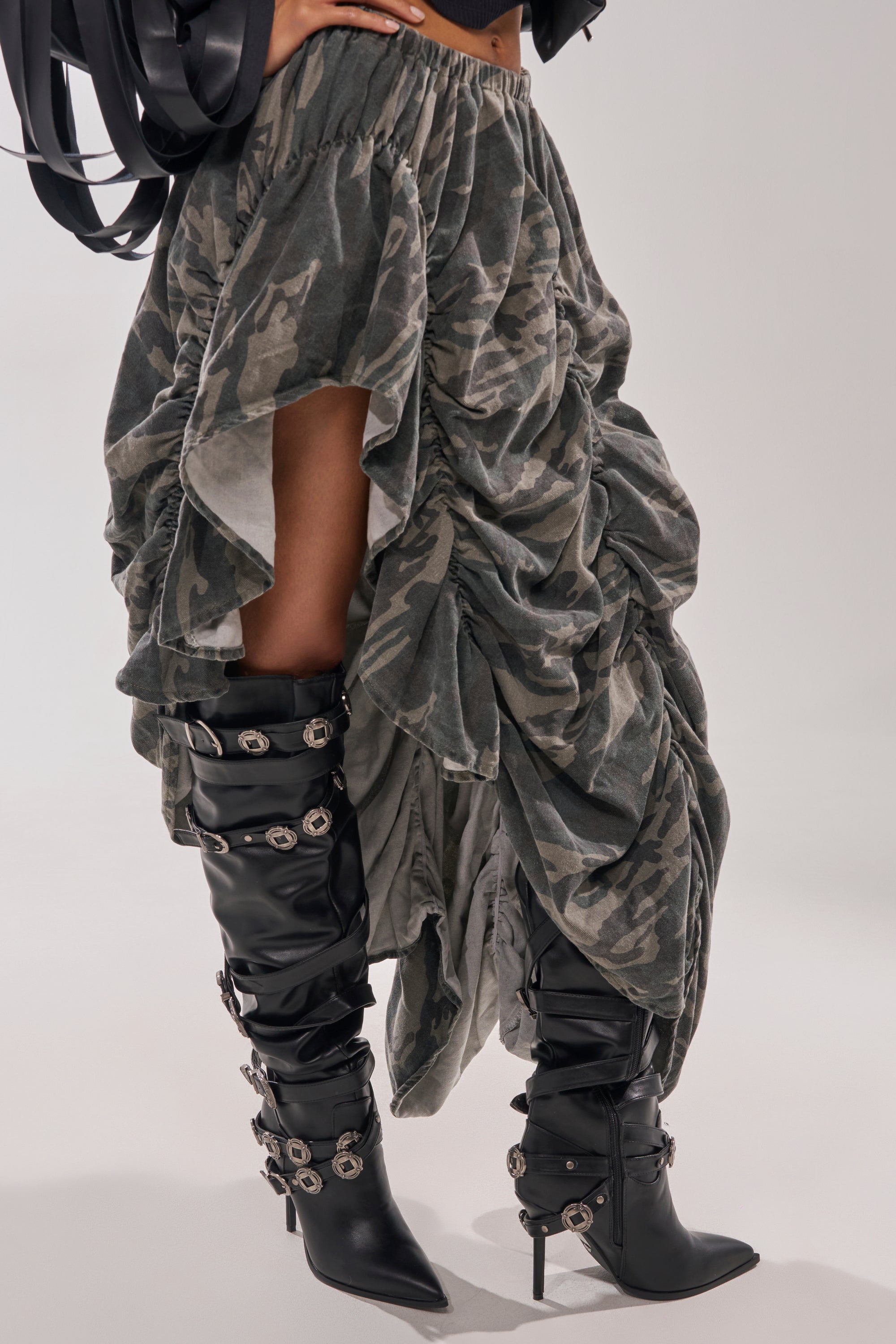 Model wears a ruched, asymmetrical camouflage skirt with AKIRA GOLD XERXES Black Stiletto Metal Embellished Boots, standing against a plain background. 2