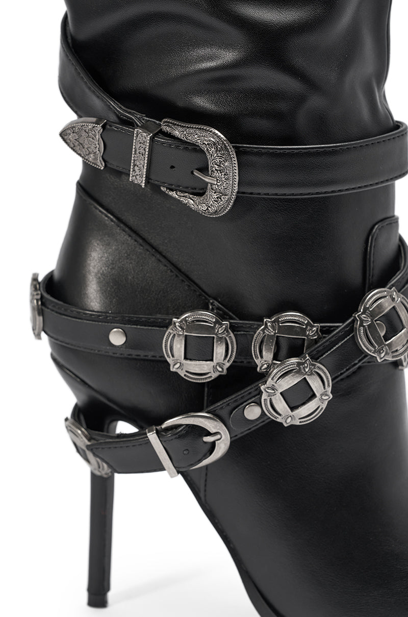 A close-up of the AKIRA GOLD XERXES BLACK STILETTO METAL EMBELLISHED BOOT in black faux leather, showcasing multiple silver buckles and straps—including a Western-style buckle near the ankle—set on a chic stiletto heel. 6