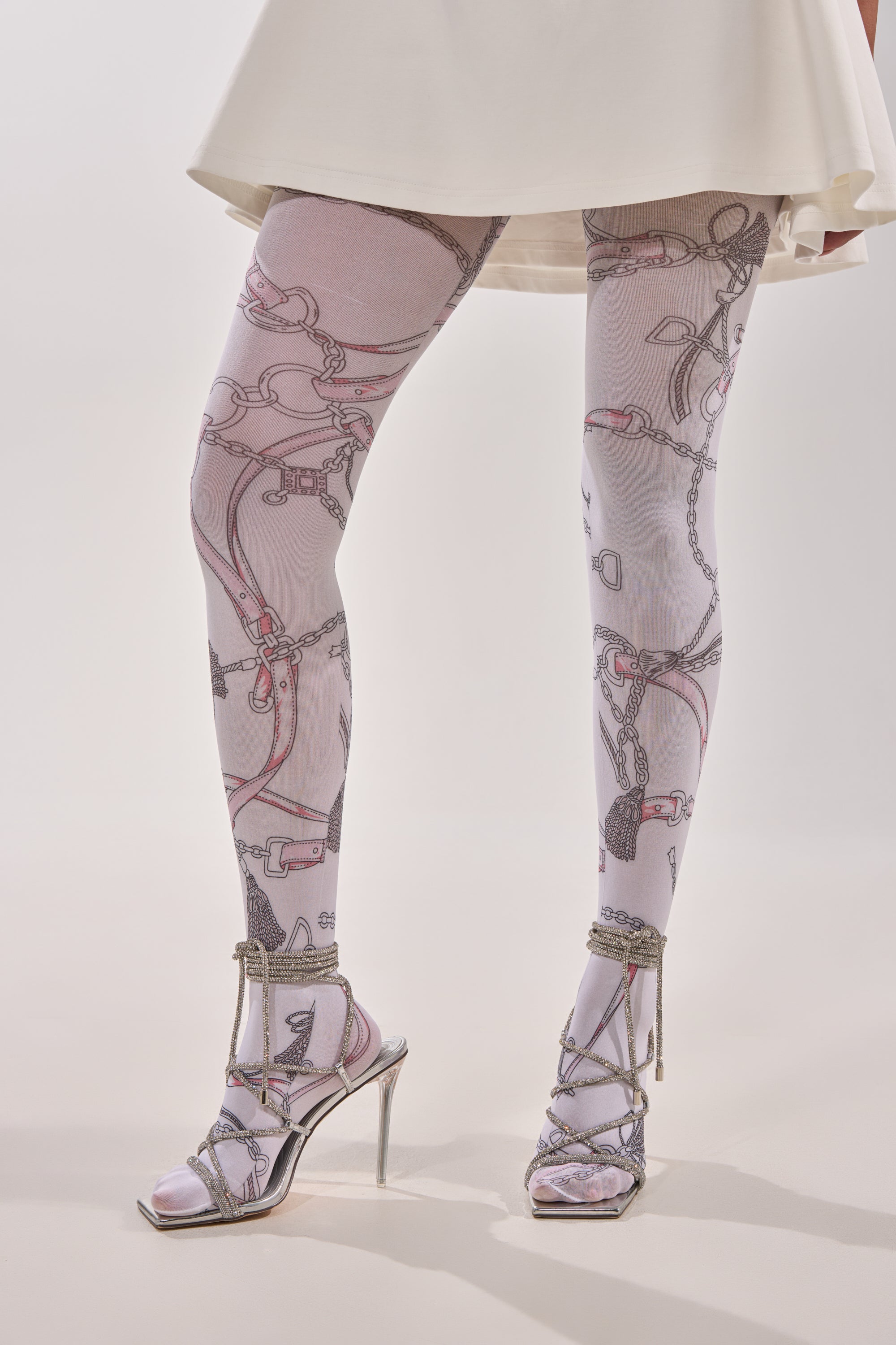 A person wears RIDE EM TIGHTS featuring a chain and strap pattern, paired with strappy silver heels and a white skirt, standing against a plain light background. 0