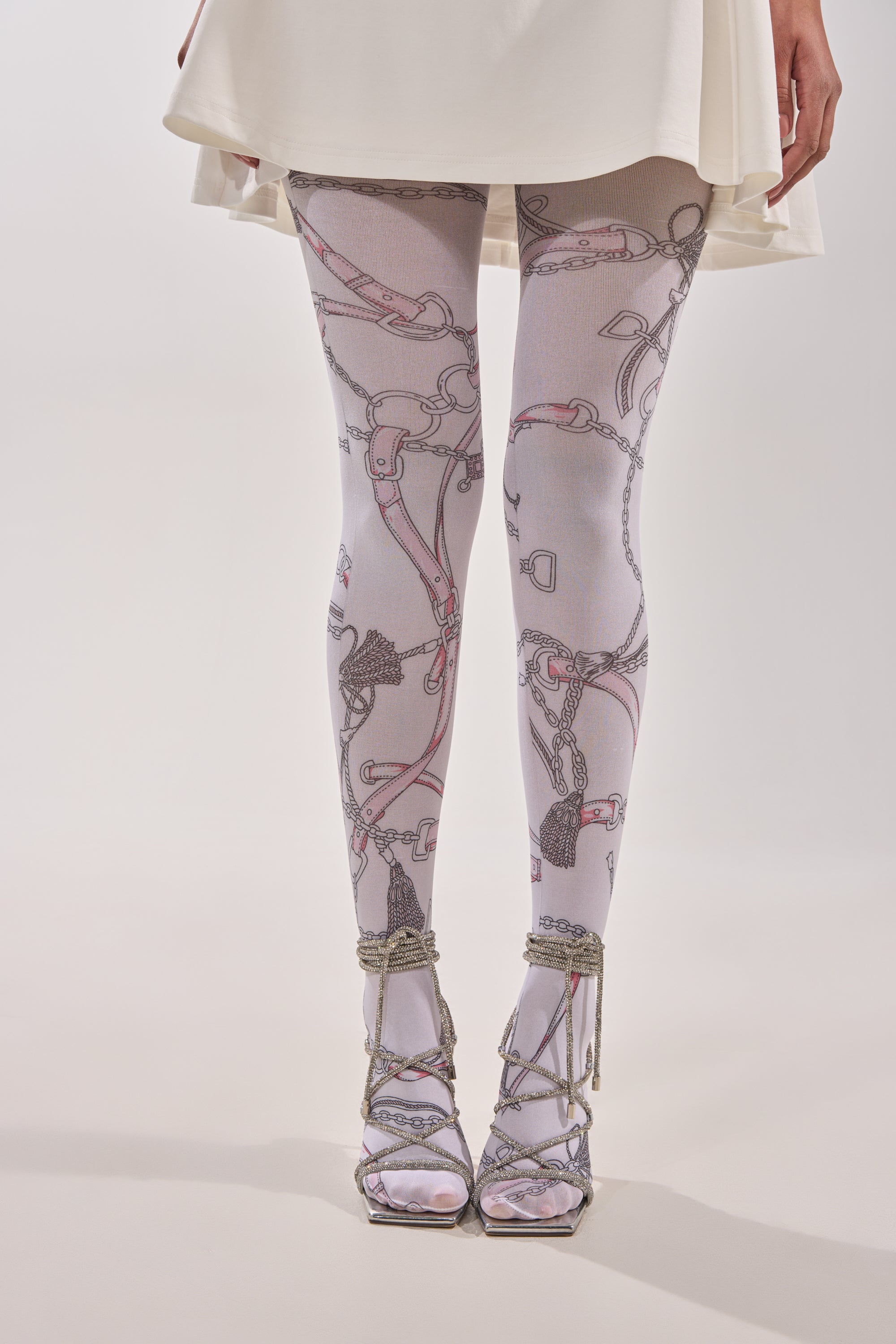 A person models RIDE EM TIGHTS—chain print tights with strap details—paired with a white skirt and silver lace-up high-heeled sandals against a plain light background. 1