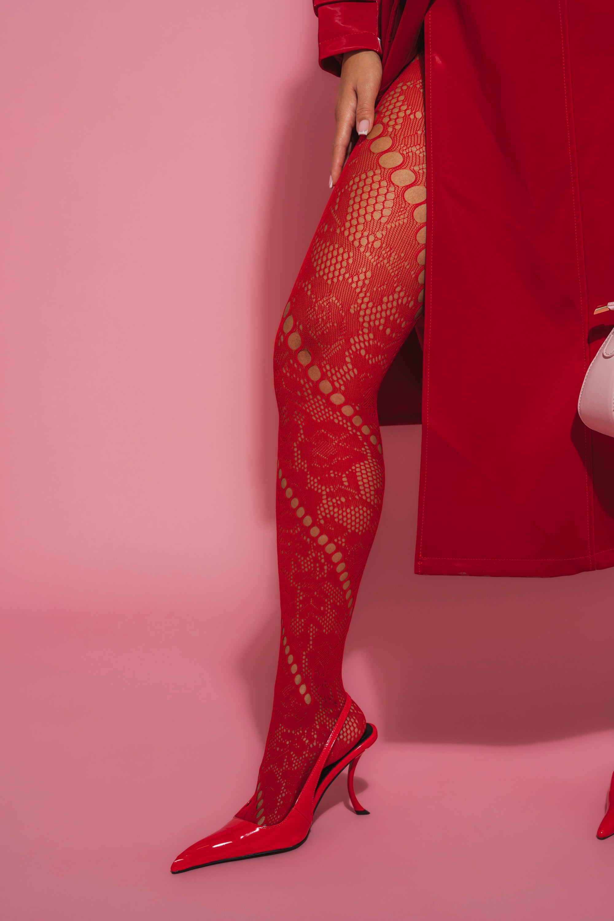 Wearing LACED FEELINGS TIGHTS, red high heels, and a red trench coat, a person stands against a pink background holding a light pink purse. 1