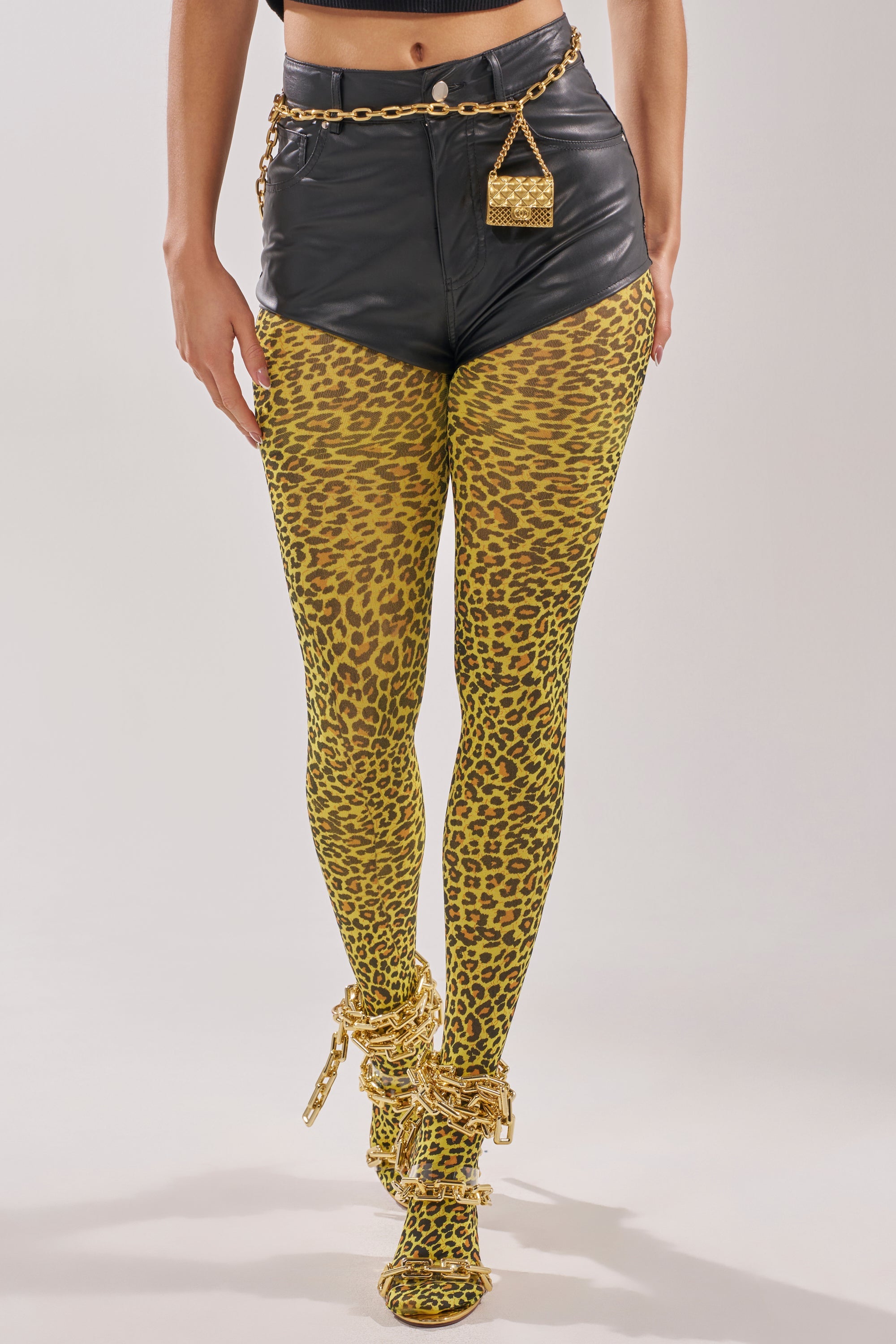 A person wears CHEETAH GURL TIGHTS—yellow cheetah print, paired with high-waisted black leather shorts, a gold chain belt with a small purse, and matching open-toe heels. The photo spotlights the lower half of the body for a bold statement look. 1