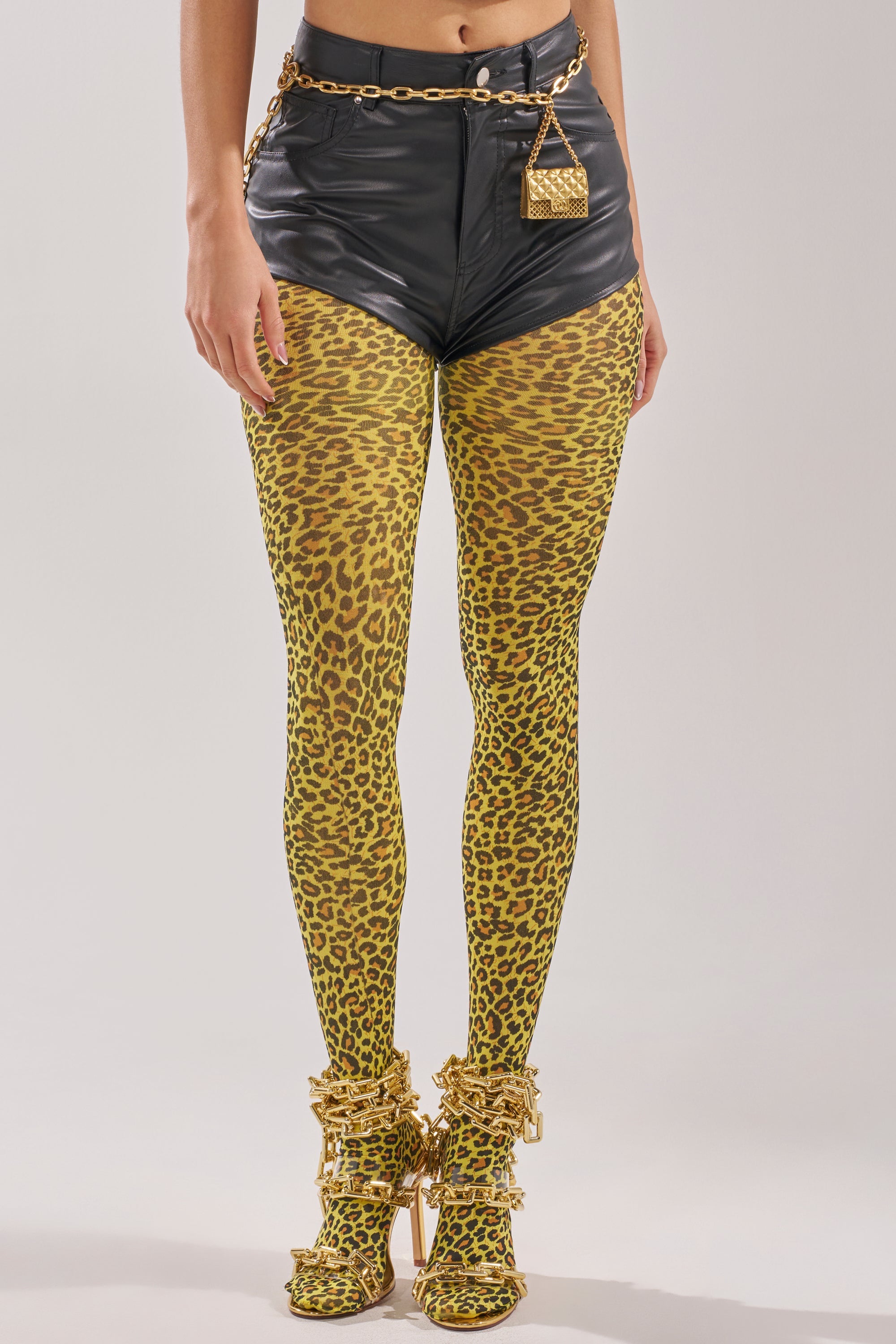 A person wearing CHEETAH GURL TIGHTS, black high-waisted shorts, a gold chain belt with a small gold purse, and gold bow-embellished heels stands against a plain light background. 2