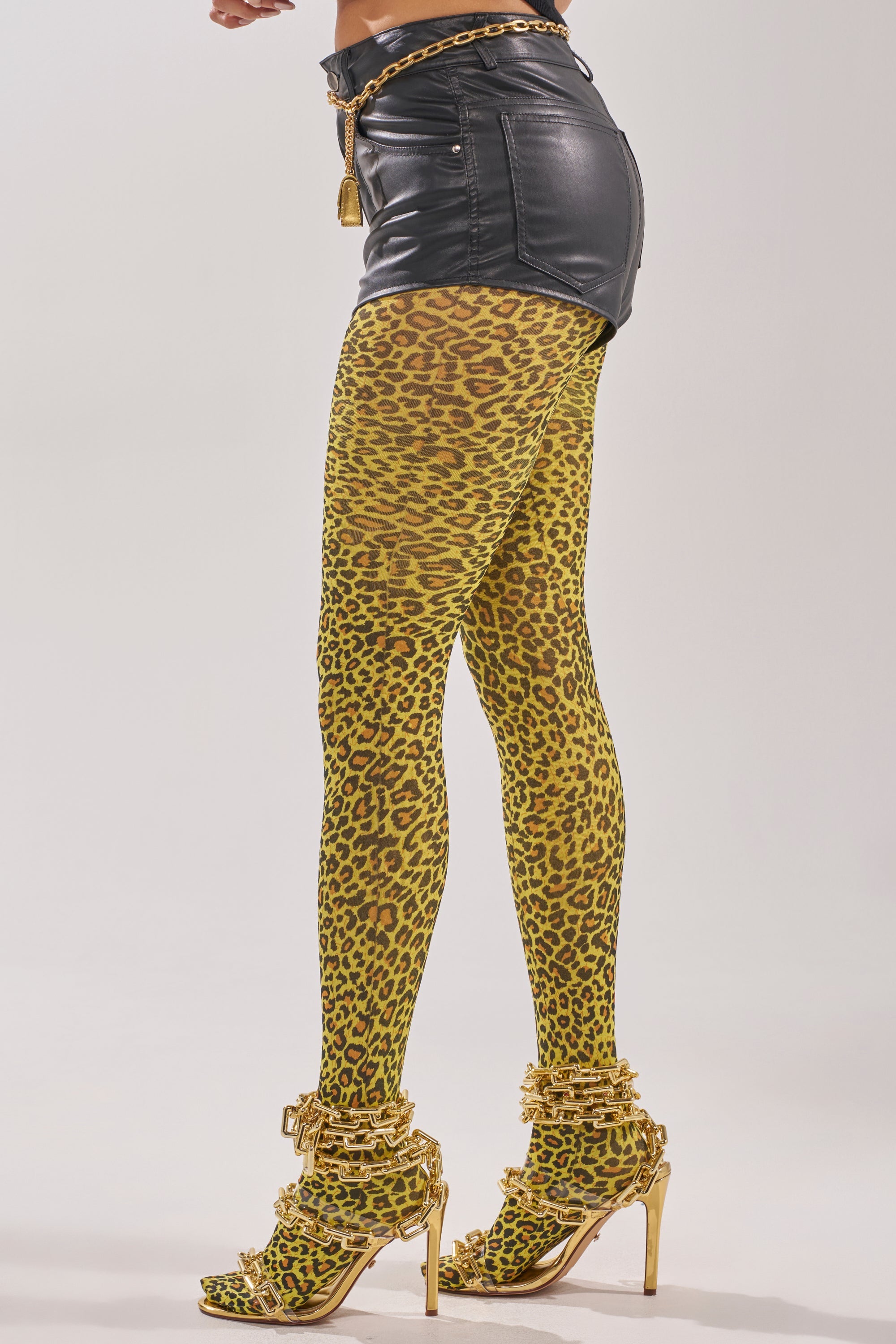 Wearing CHEETAH GURL TIGHTS with black leather shorts and gold high-heeled sandals adorned with gold accessories, a person stands against a plain light background. 3