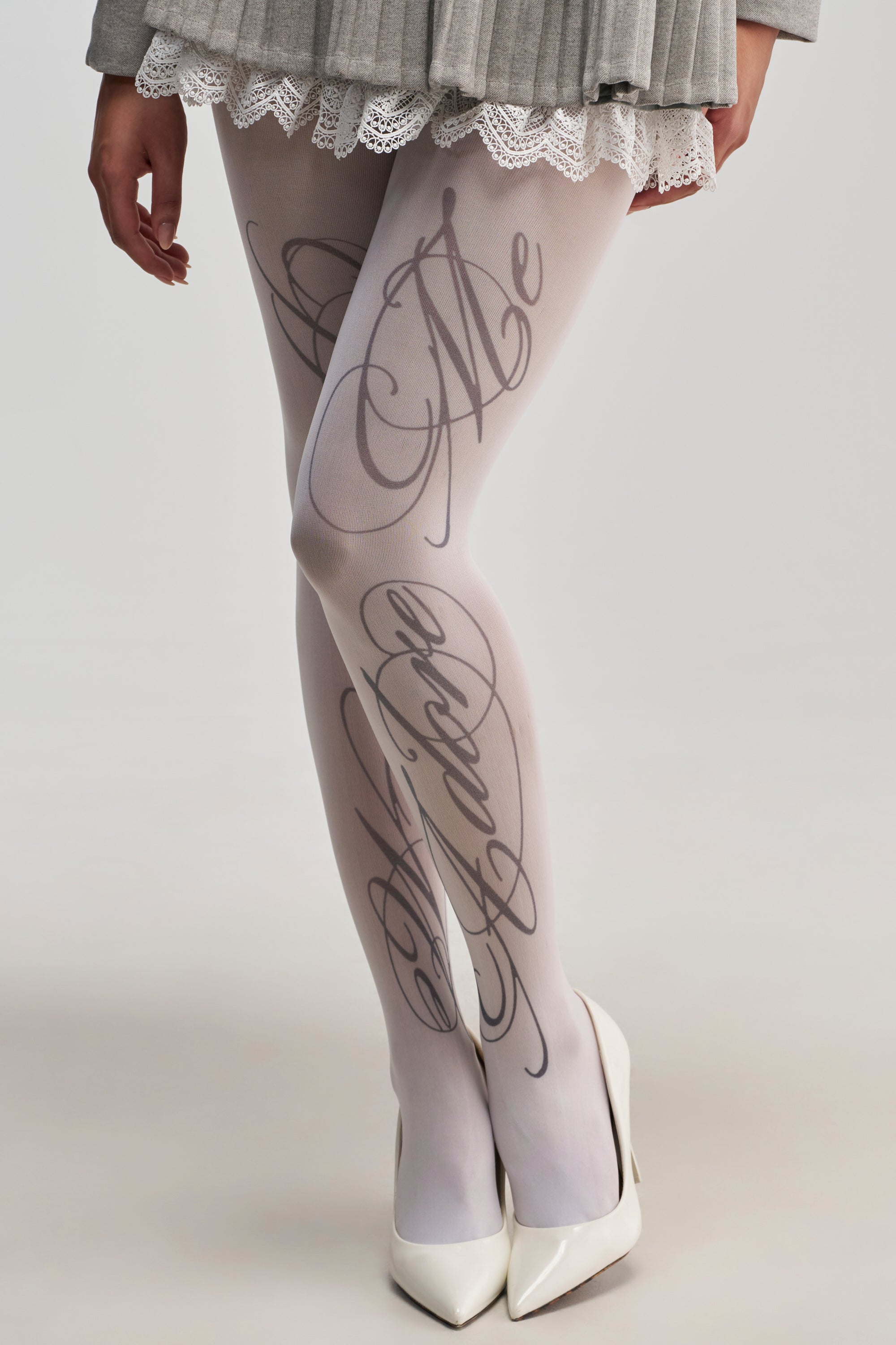 A person wearing white high heels, a grey pleated skirt with lace trim, and the I DO GRAPHIC TIGHTS—white tights featuring elegant cursive lettering. The image highlights the legs and lower part of the skirt against a neutral background, showcasing chic streetwear accessories in a minimalist setting.
