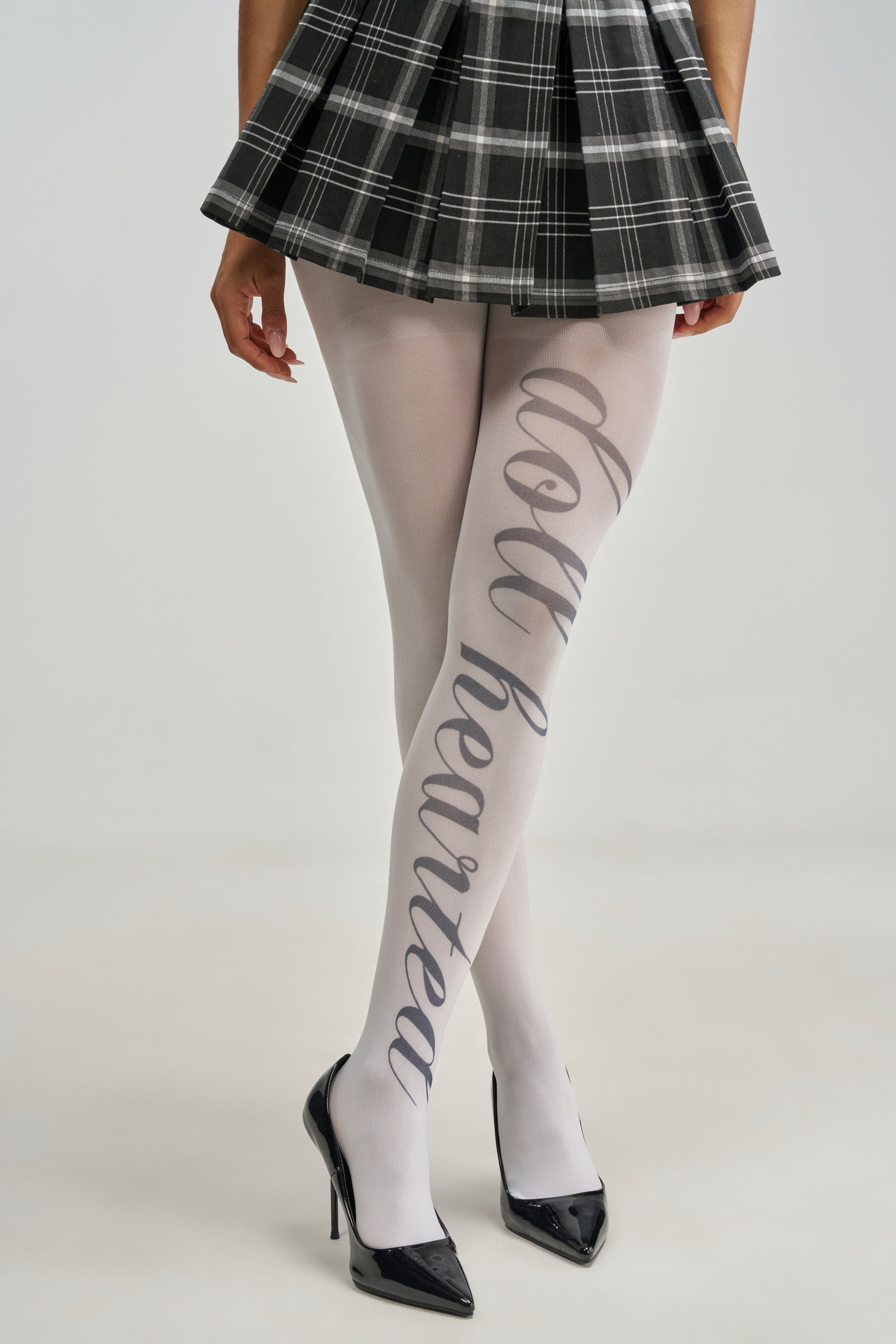 HEY DOLL GRAPHIC TIGHTS in white mesh with “dollhearted” printed vertically up the left leg, worn with a pleated plaid skirt and black stiletto heels.