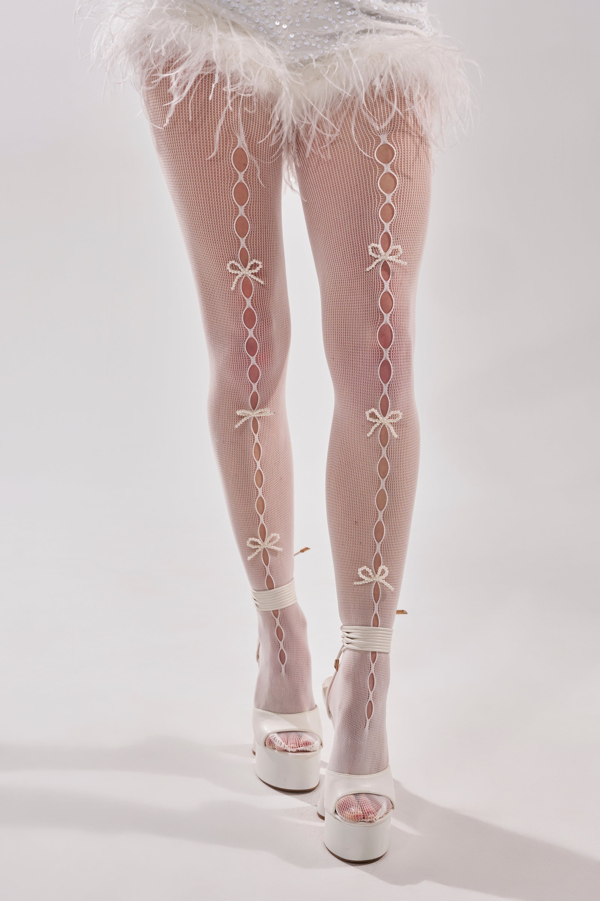 Wearing ANGEL SEASON TIGHTS featuring a decorative pearl bow pattern down the back, paired with white platform heels and a feather-trimmed skirt, a person stands against a light background. 2