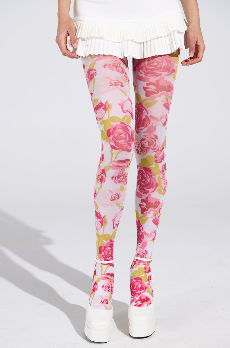 A person wears the STYLE ME UP ROSE TIGHTS with pink roses and green leaves, paired with white platform shoes and a short pleated skirt. The high-rise fit enhances floral elegance against a plain light gray backdrop.