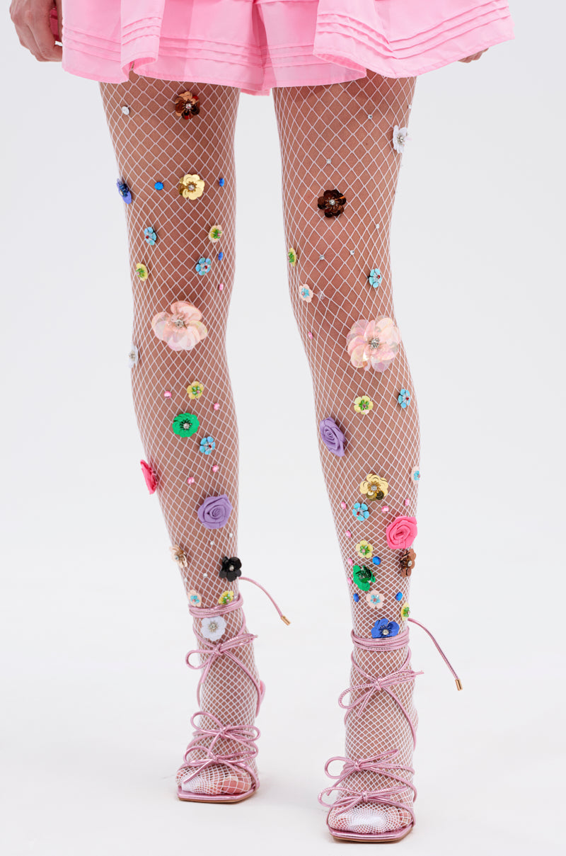 A person stands against a plain light background wearing ITS POPPIN EMBELLISHED TIGHTS—white tights with colorful floral embellishments—paired with a pink skirt and pink lace-up sandals.