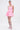 A woman stands against a plain background wearing ITS POPPIN EMBELLISHED TIGHTS—stretchy white fishnet tights with floral details—paired with a pink dress, matching heels, pink earrings, and her hair tied back.