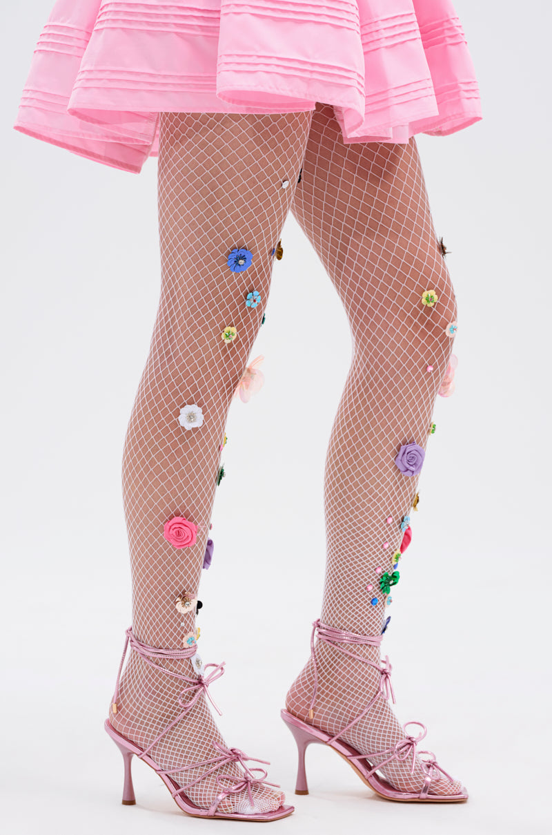 Model wears the ITS POPPIN EMBELLISHED TIGHTS—white fishnet tights with vibrant floral accents—paired with a pink skirt and strappy pink heels, shown waist down against a plain background.
