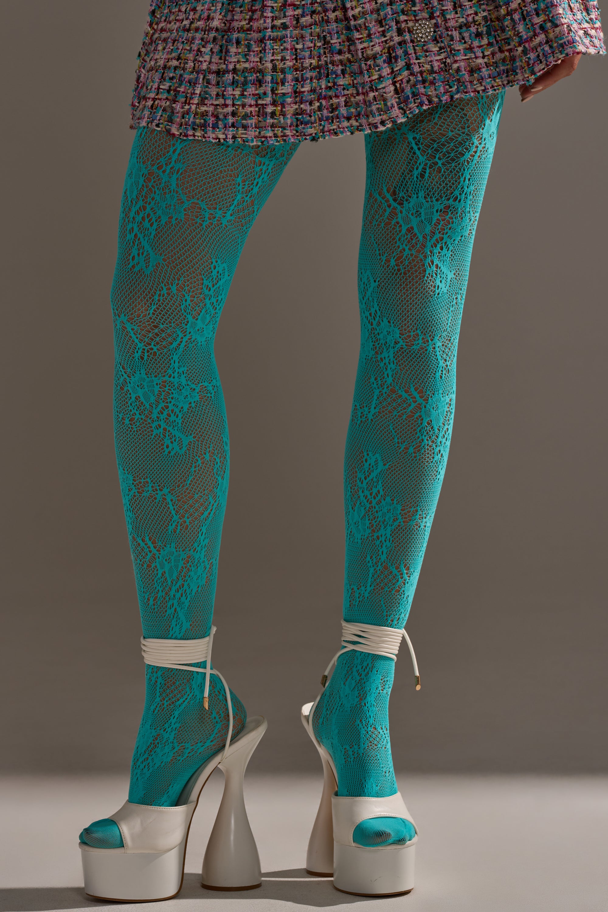 Wearing LUXE LIVING TIGHTS in an aqua-blue lace pattern, a multicolored textured skirt, and white ankle-strap platform high-heel sandals, the person stands against a plain background.