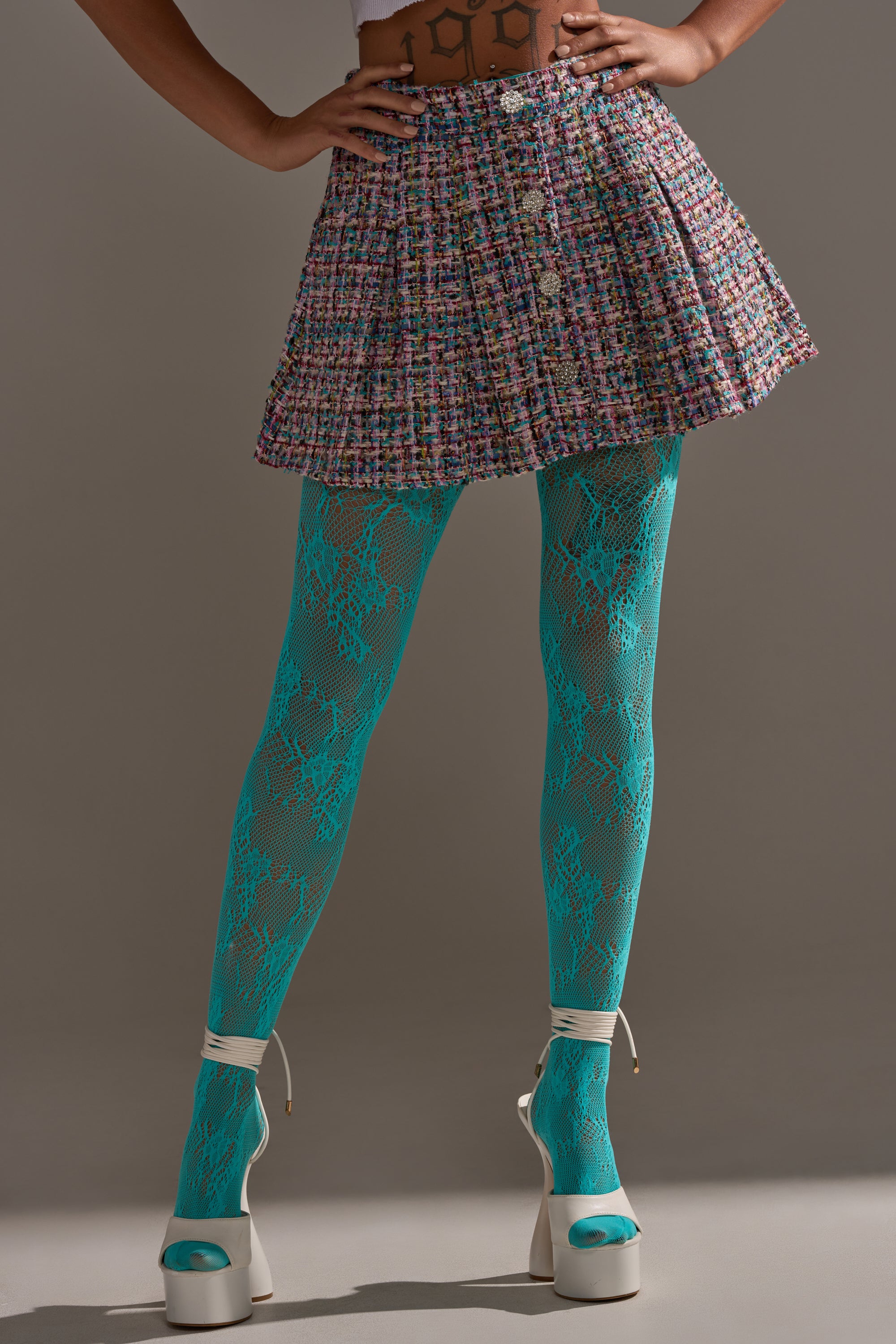 Model wears a pleated multicolored tweed skirt, LUXE LIVING TIGHTS in lacy aqua-blue, and white platform high heels, standing with hands on hips against a plain backdrop.