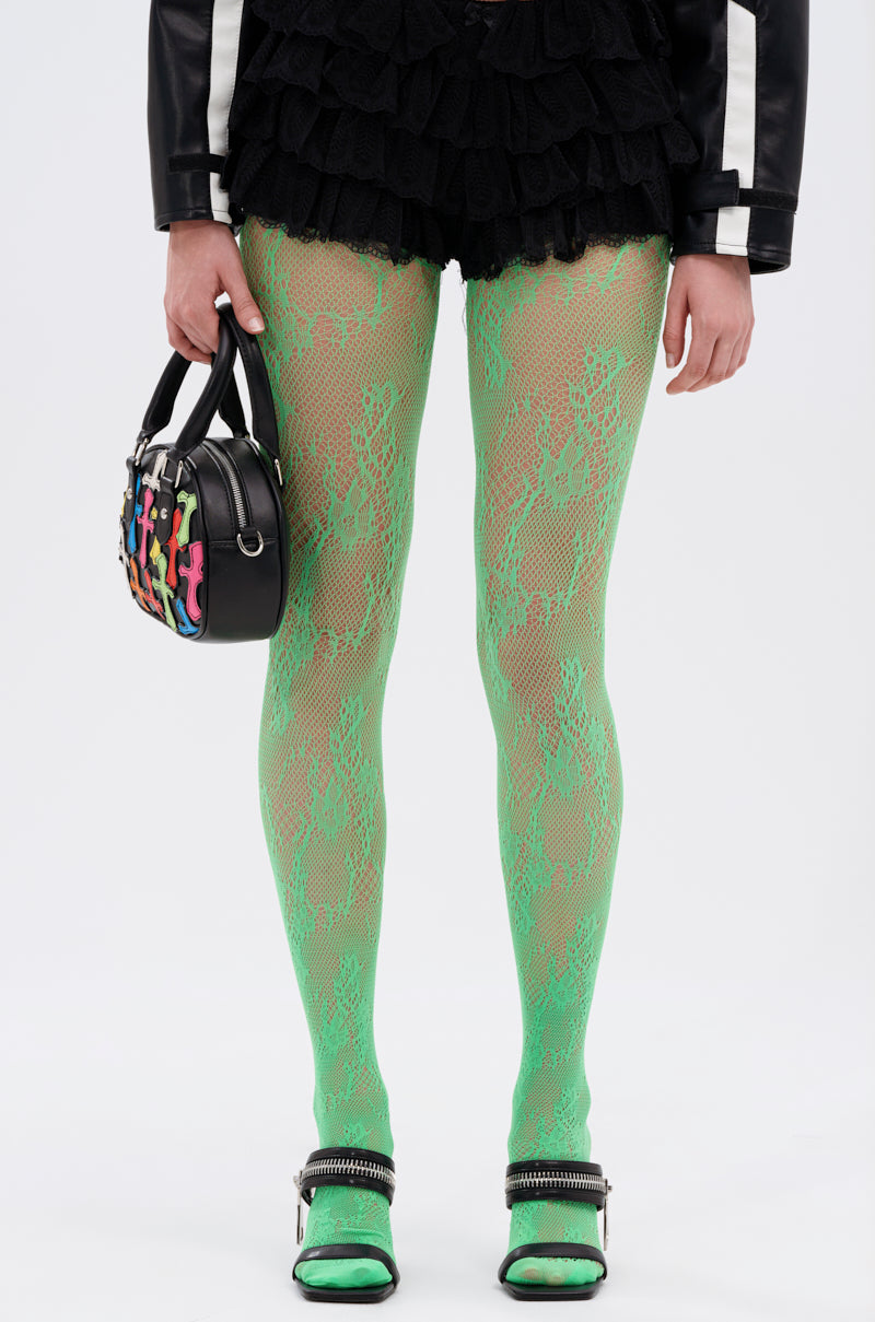 A person wearing GREEN WITH NO ENVY TIGHTS, a black high-rise ruffled skirt, a black jacket with white sleeve stripes, silver-embellished black shoes, and holding a small black handbag with colorful accents.