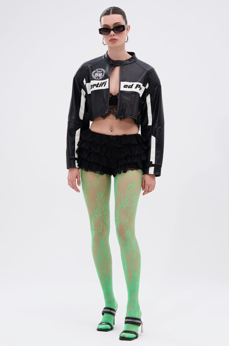 A person wears GREEN WITH NO ENVY TIGHTS, a cropped black leather jacket, a black high-rise ruffled mini skirt, open-toe heels, black sunglasses, and has slicked-back hair against a plain white background.