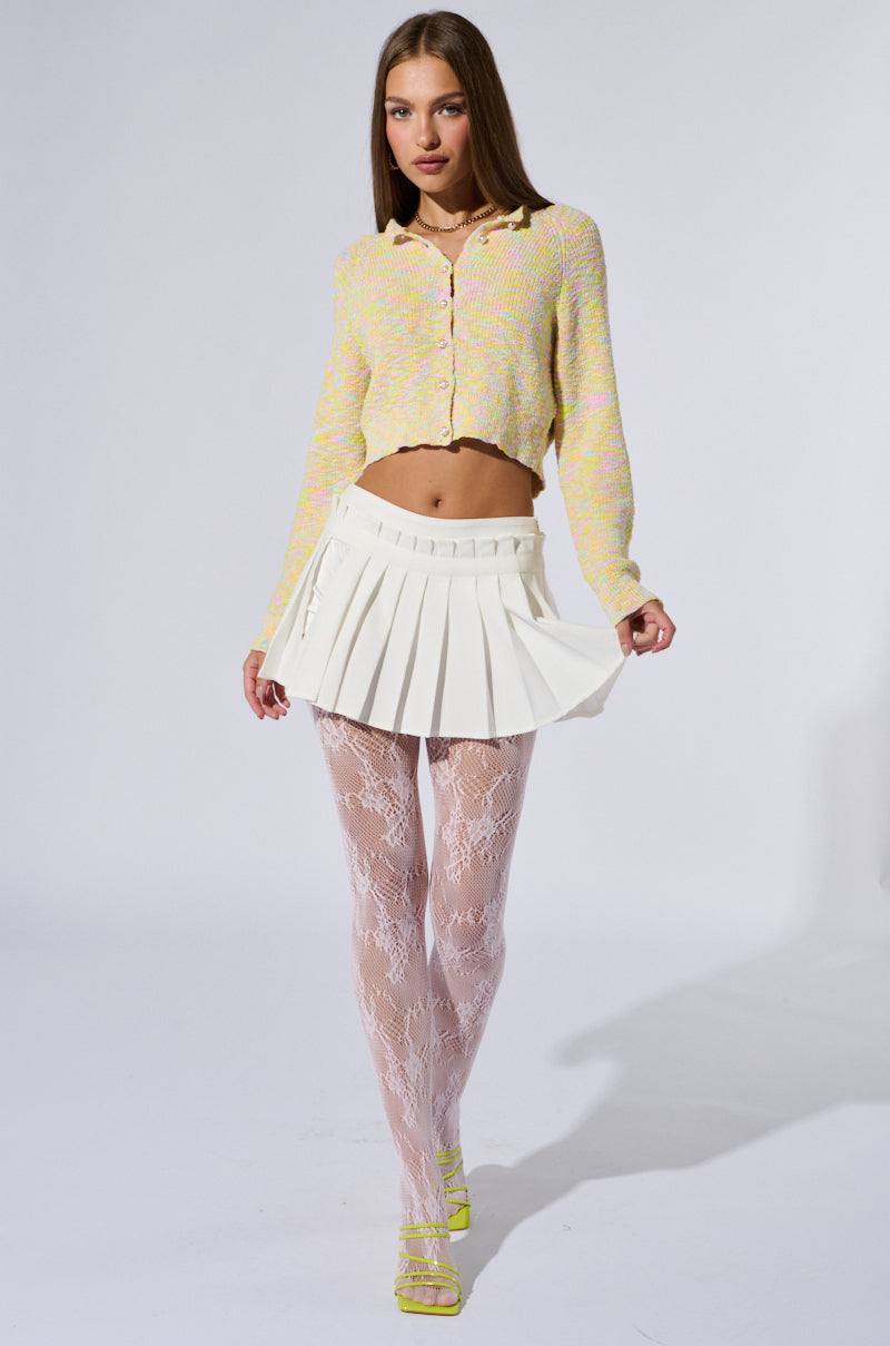 A person models a stylish outfit consisting of a pastel yellow cardigan, a white pleated mini skirt, PRETTY PINK LACE TIGHTS adorned with a dainty lace pattern, and strappy yellow heels. They are posing against a plain white background.