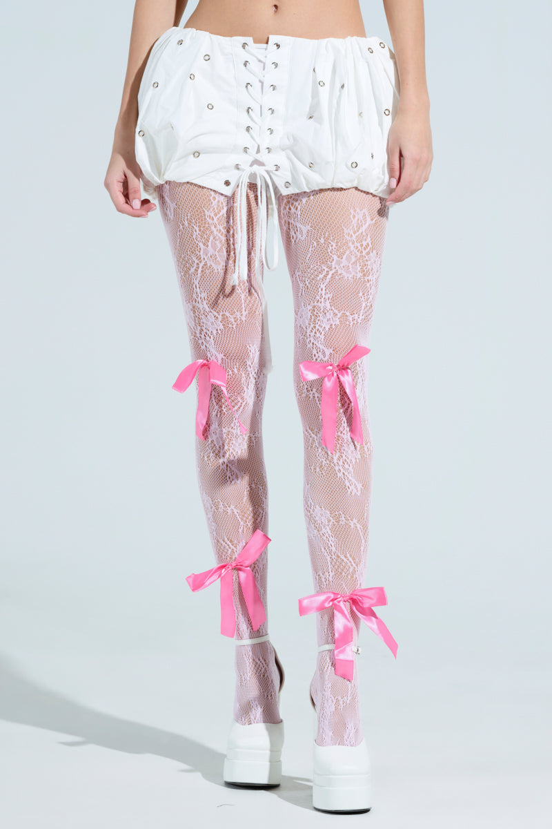 Someone dons a white corset-style skirt paired with PRETTY IN PINK BOW TIGHTS, featuring bubblegum pink nylon lace with satin bow appliques. White platform shoes complete the ensemble.