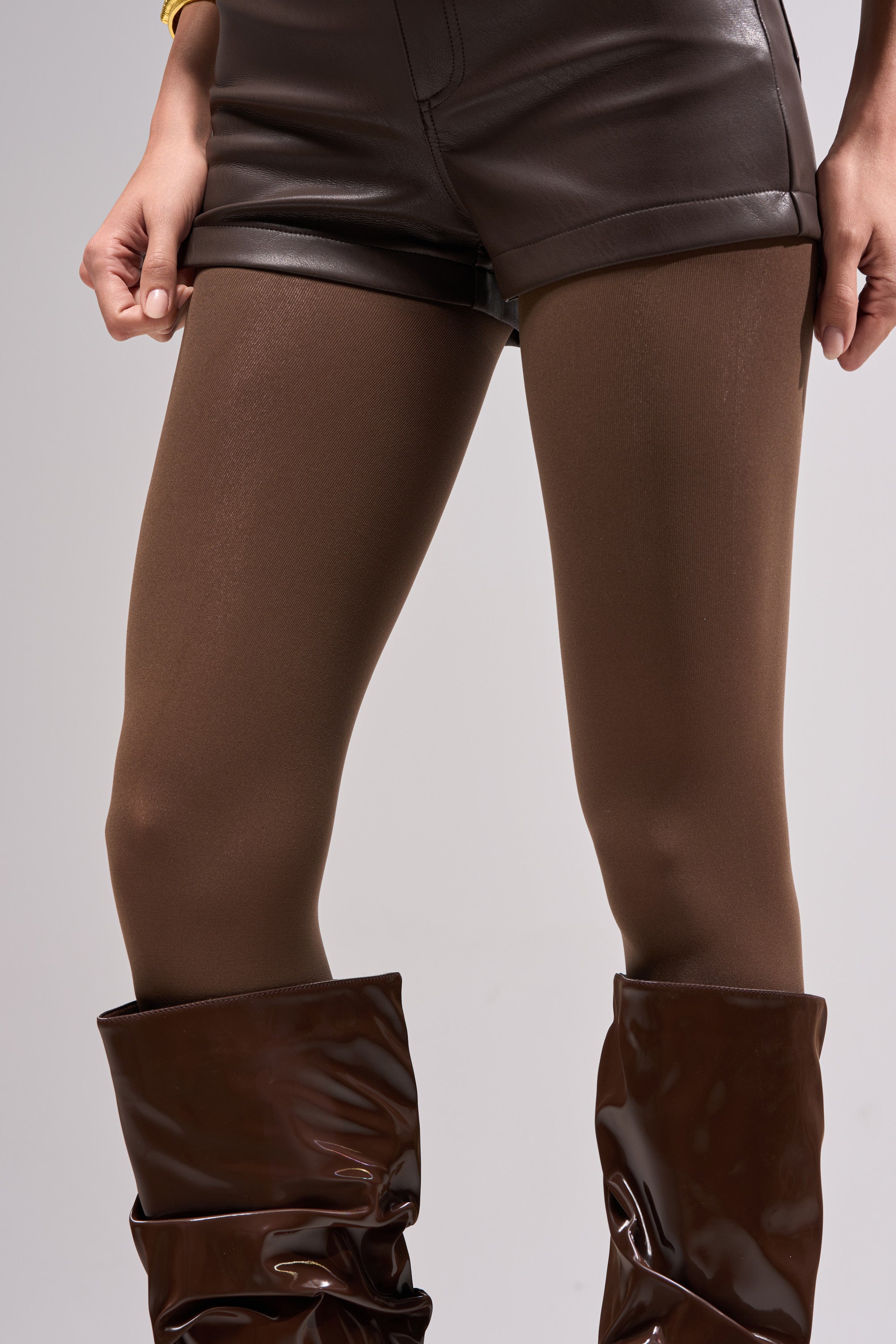 A person stands with hands at their sides in CHOCOLATE NUDE TIGHTS, glossy brown shorts, and shiny brown knee-high boots against a light gray background.